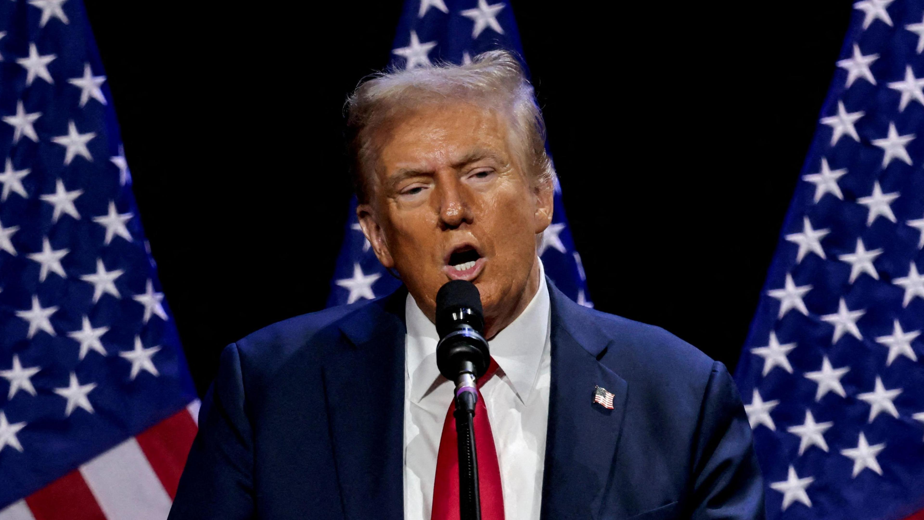 An image of Donald Trump, a man with short light hair wearing a blue suit, white shirt and red tie, speaking into a microphone standing in front of three US flags and a black background