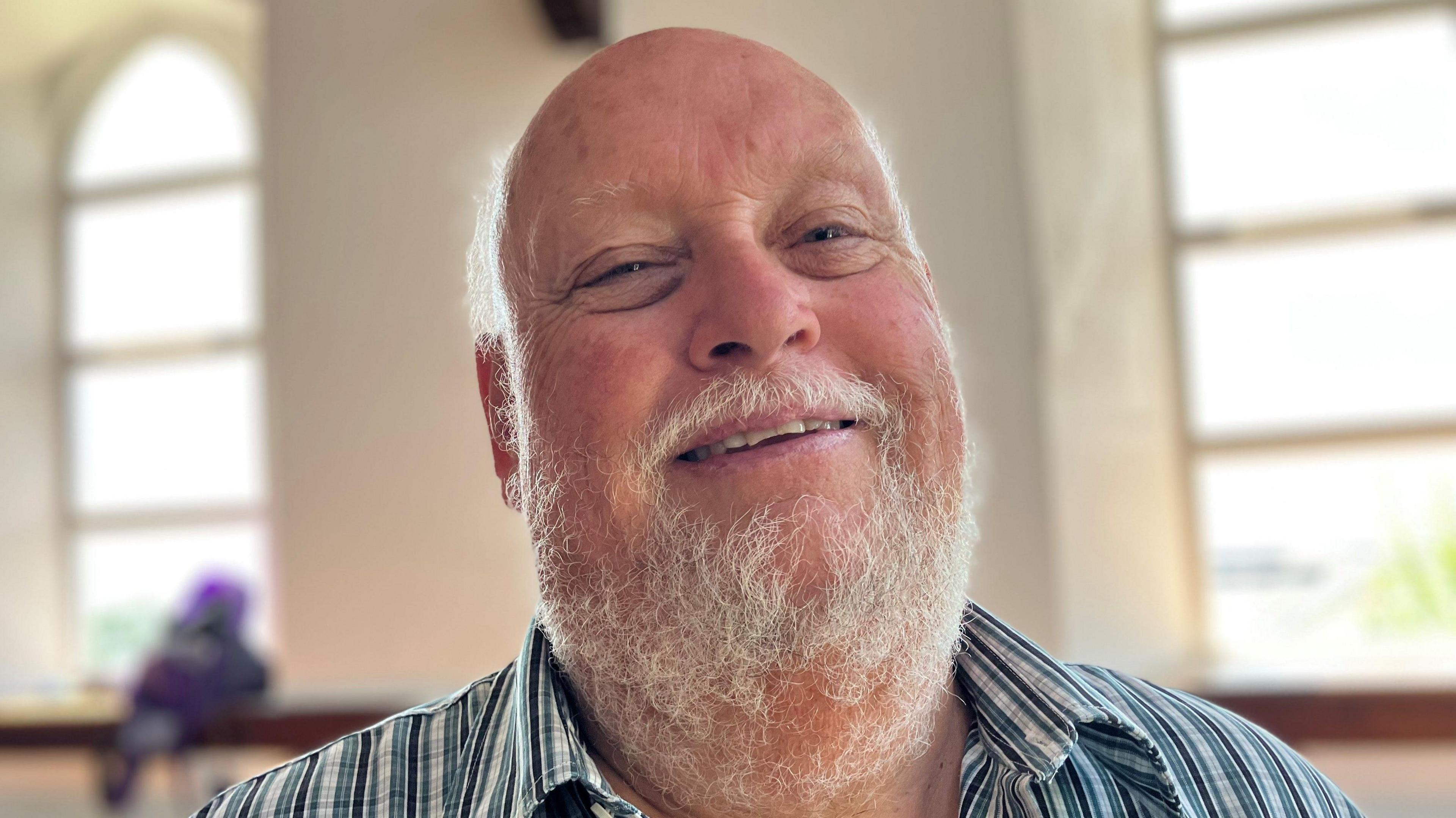 Bald man with a white beard and moustache smiling at the camera with the gothic chapel windows behind
