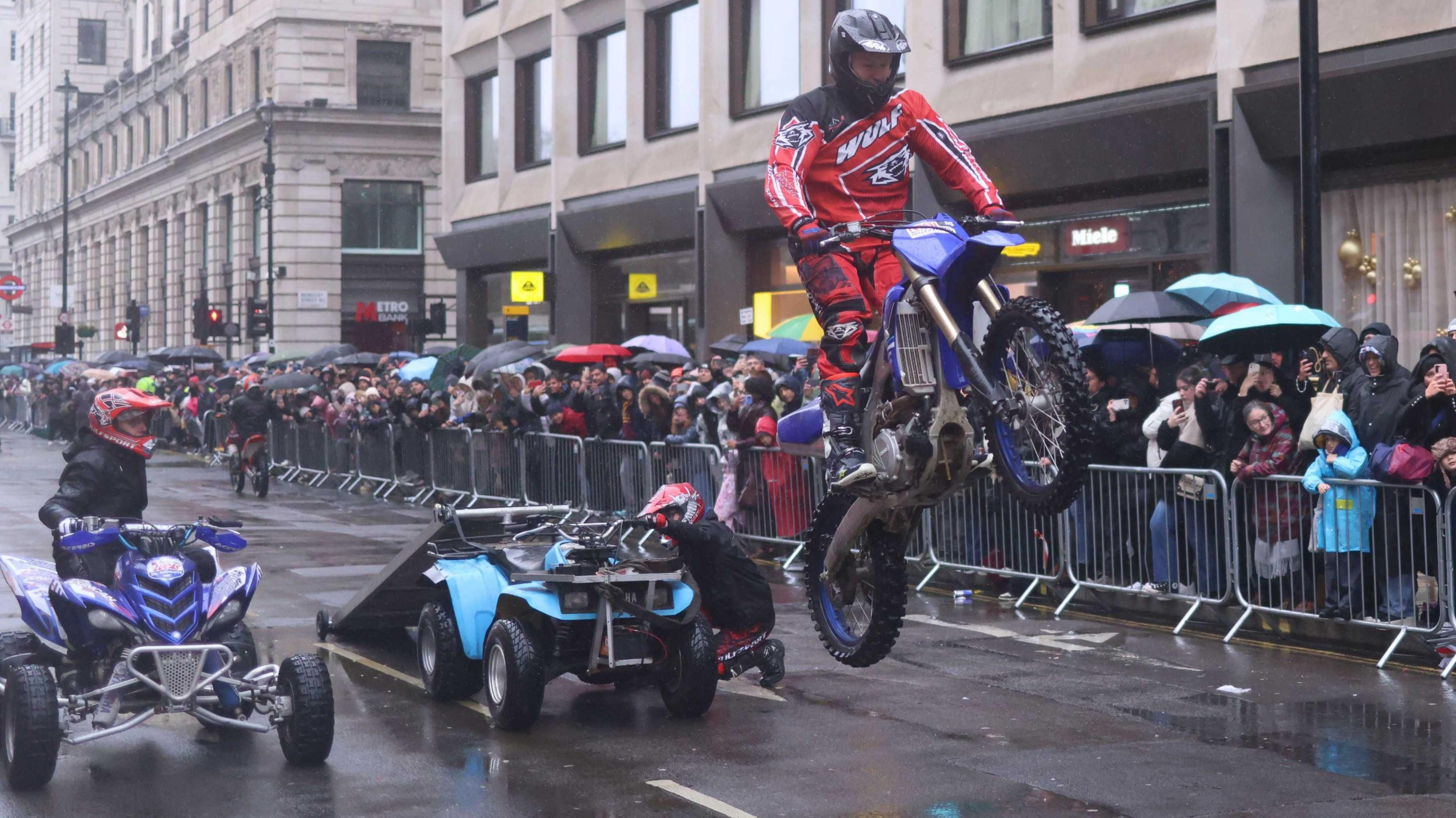 A man on a motorbike, wearing a helmet, pulls a wheelie as a crowd of people watches. Behind him are two quad bikes. 