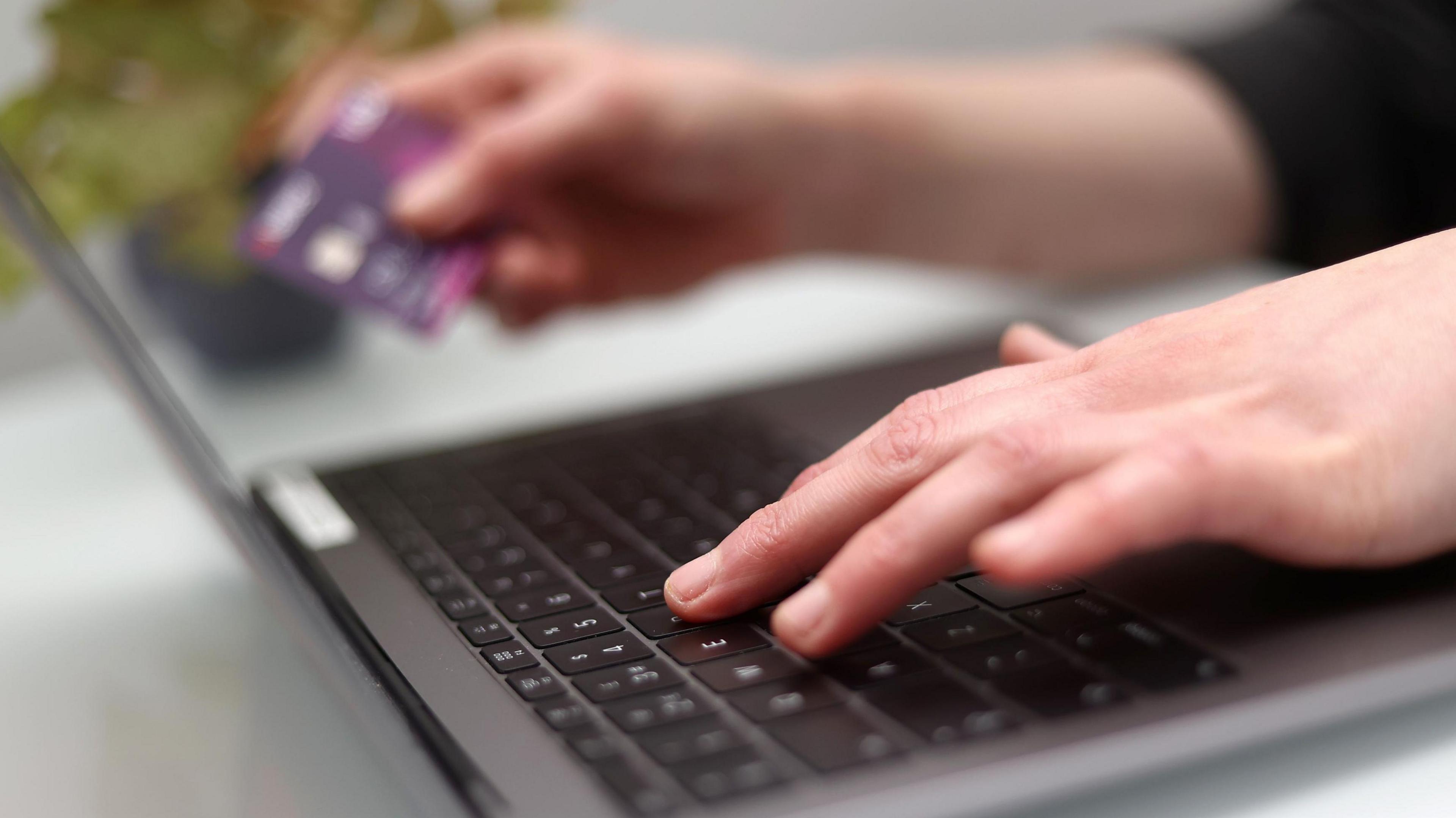 A woman uses a laptop as she holds a bank card. Her fingers can be seen typing on a black keyboard. 