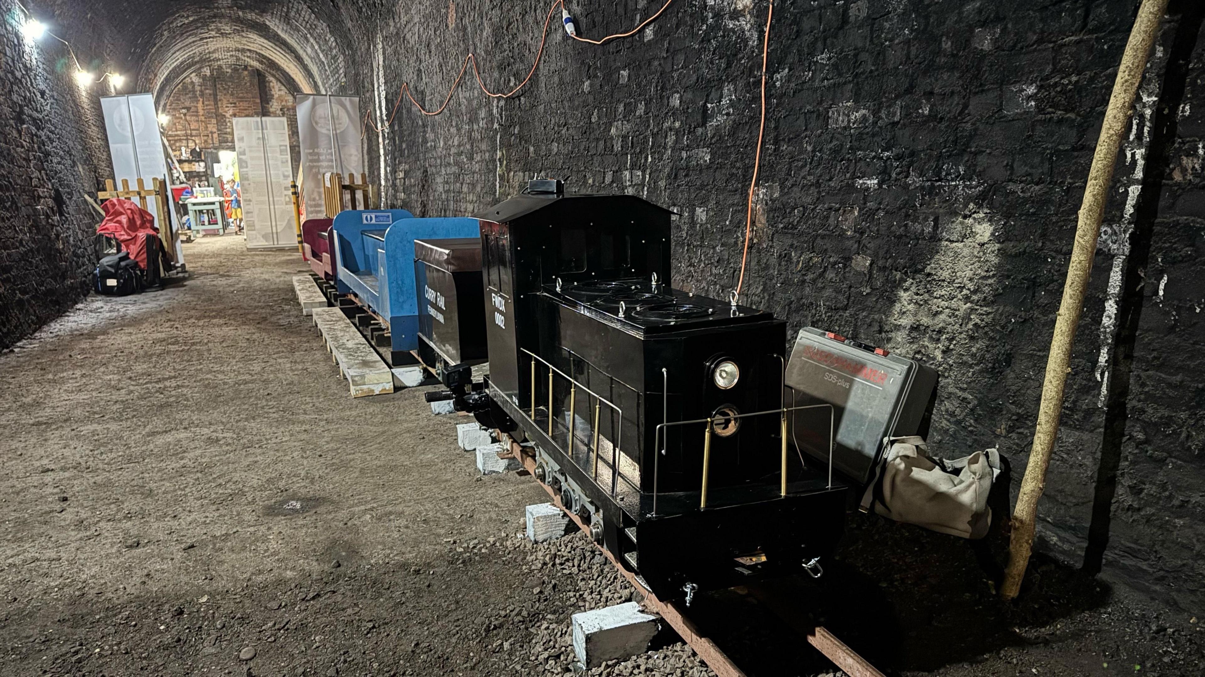 A miniature railway on small tracks in an arch-shaped tunnel with dark brick walls. The front of the train is back with silver railings round the side.