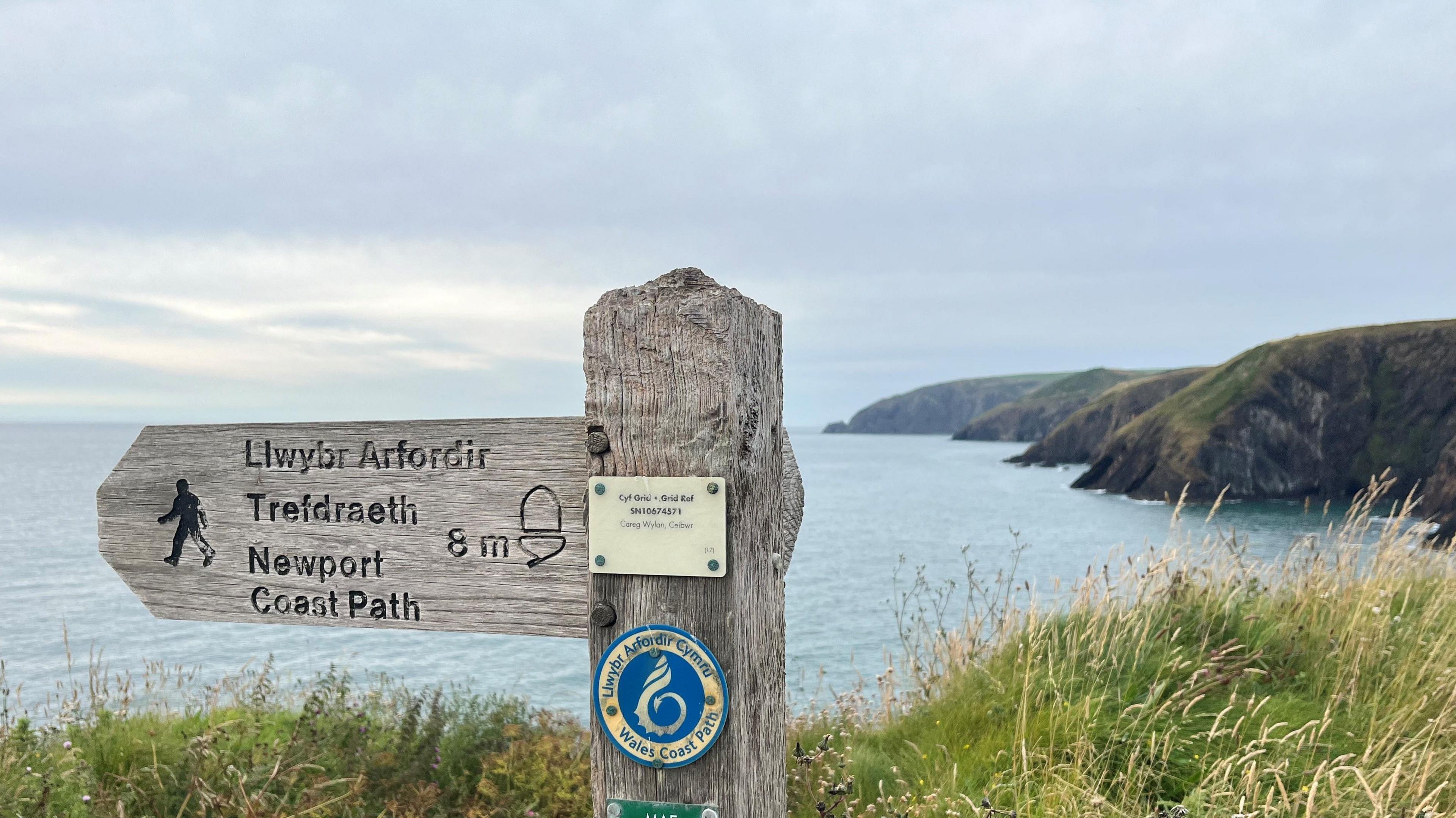 Photo of Ceibwr Bay, a public footpath sign is in the foreground, with the words Llwybr Arfordir, Trefdraeth and Newport Coast Path written alongside an acorn image. In the background is grass, the sea and four headlands. 