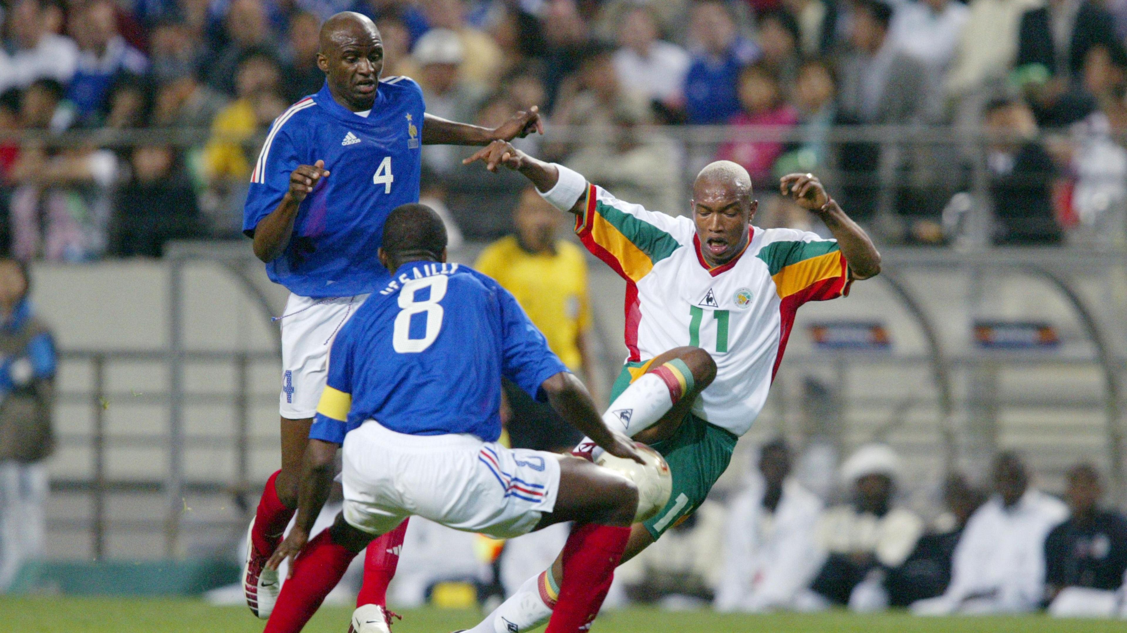 El Hadji Diouf, wearing a predminantly white Senegal kit trimmed with the green, red and yellow colours of the national flag, raises his arms to balance as he looks to swivel away as he controls a football, while France team-mates Patrick Vieira and Marcel Desailly, wearing blue shirts, white shorts and red socks, attempt to dispossess him. Blurred in the background is a dugout with people sitting in it and fans in a stand