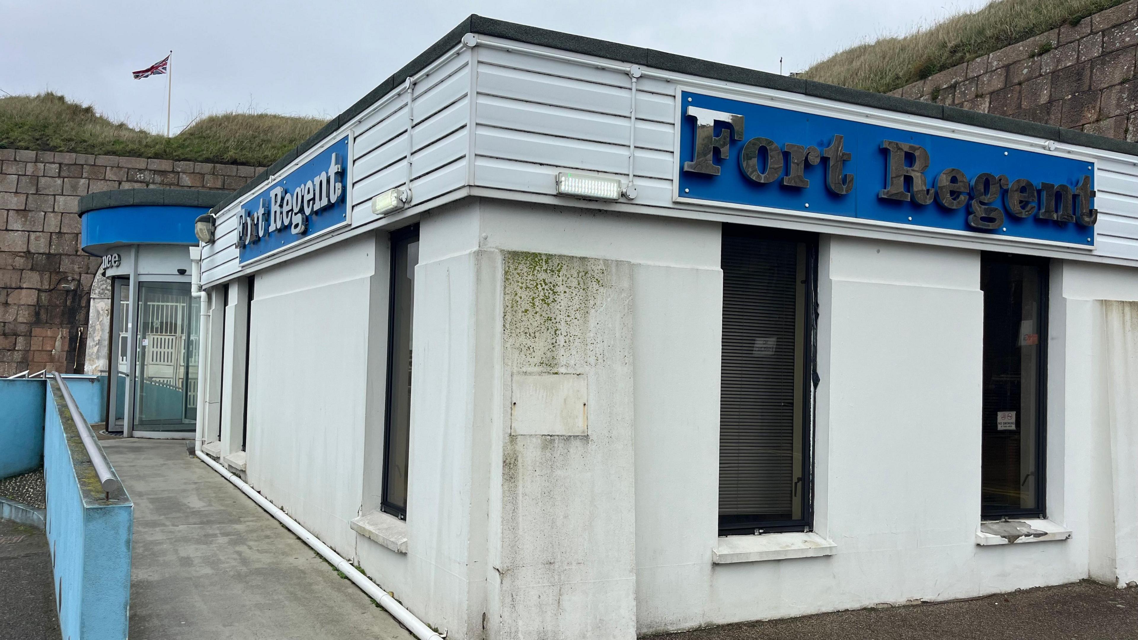 The front of the Fort Regent building. It is white with blue features and silver letters spelling out Fort Regent.