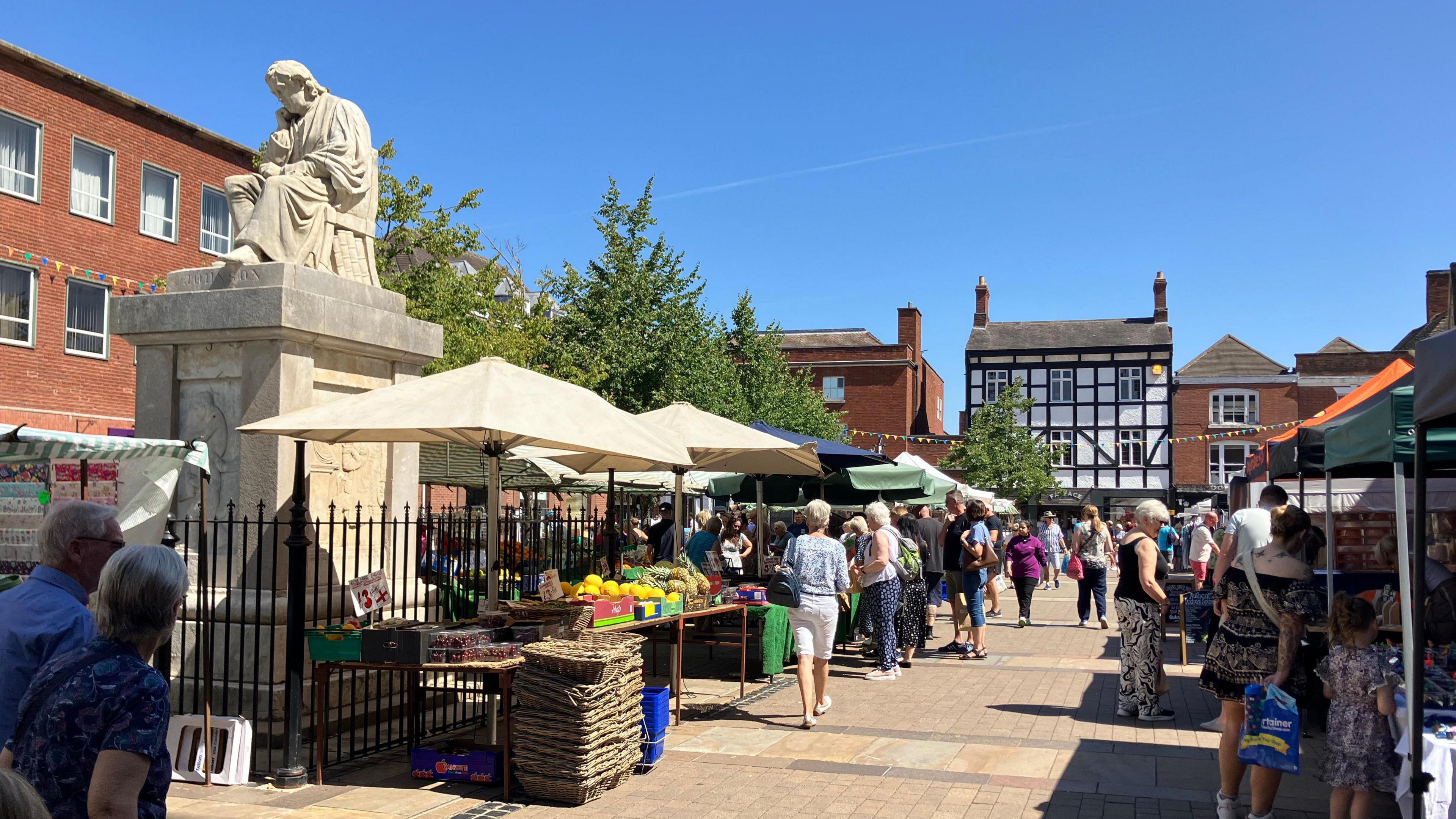 Lichfield town centre, with market stalls set up and people milling around. There is a clear blue sky, and a number of buildings surround the area where the market is being held.