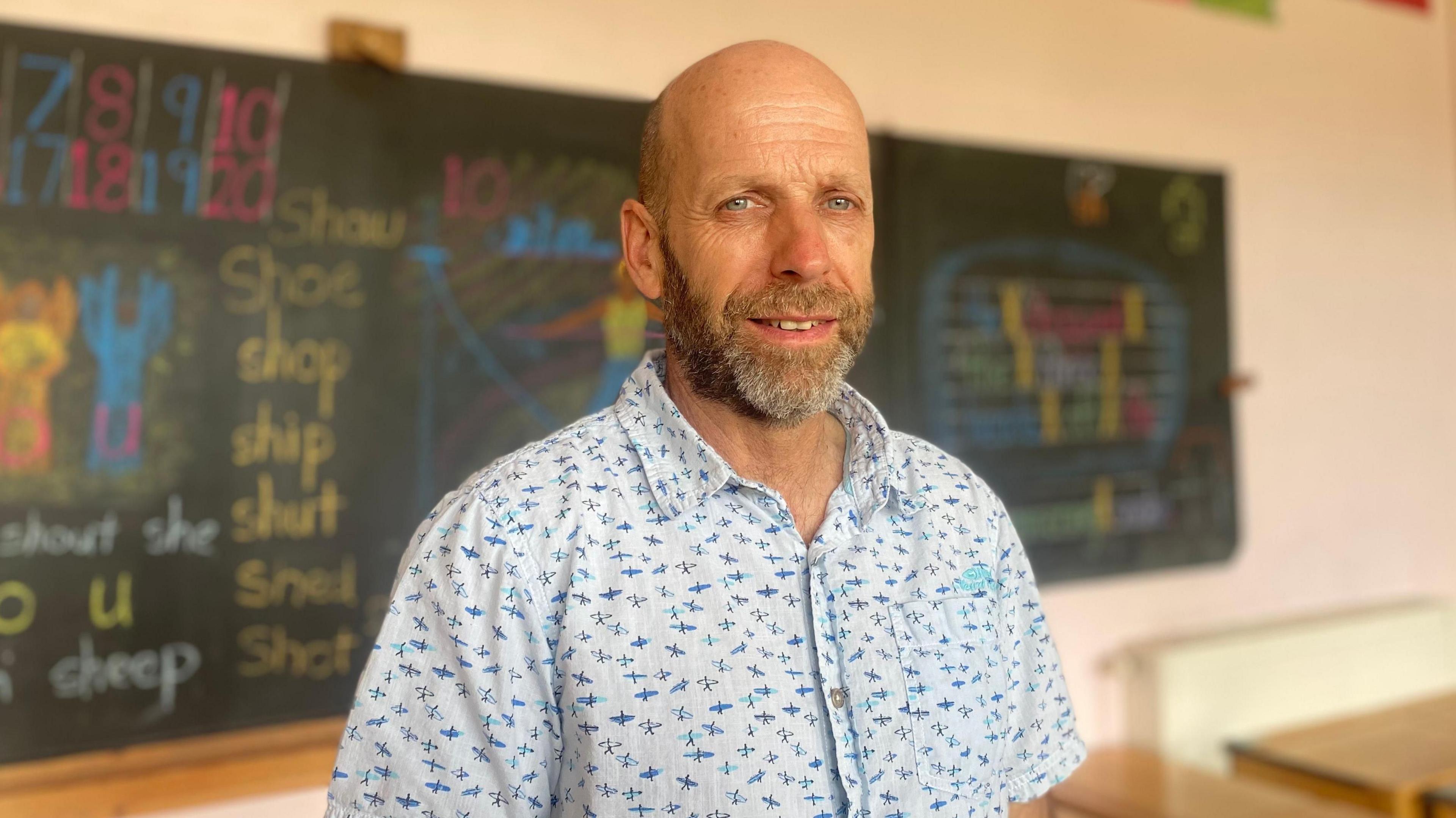 Head teacher Rob Strafford stood in front of a chalkboard and wearing a short-sleeved shirt, with a beard and smiling at the camera