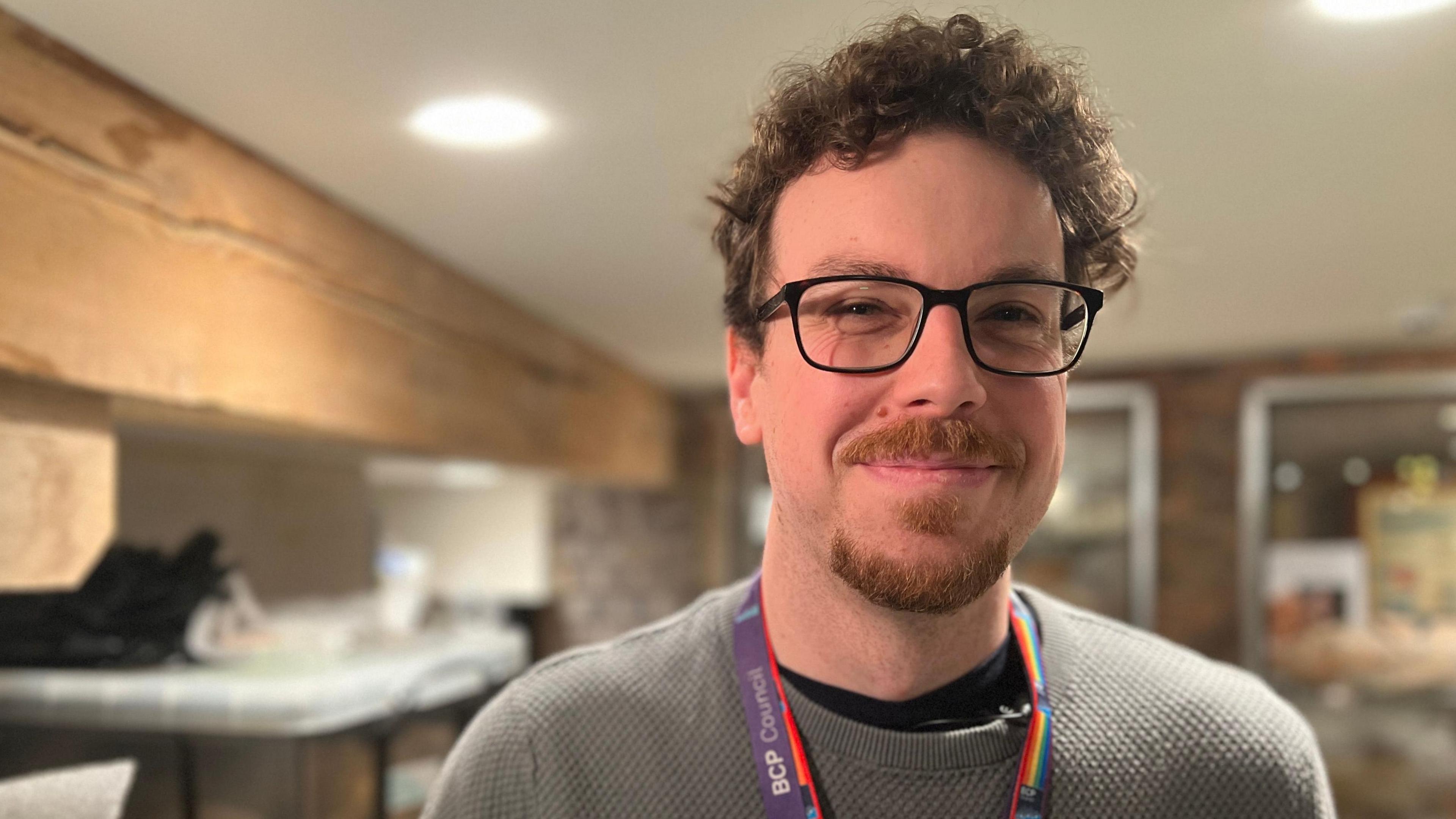 Joe Raine wears a grey jumper and rainbow coloured lanyard that reads "BCP council". He is standing in the collections room of Poole Museum. The background is out of focus but some plastic boxes containing finds can be discerned in the background. 