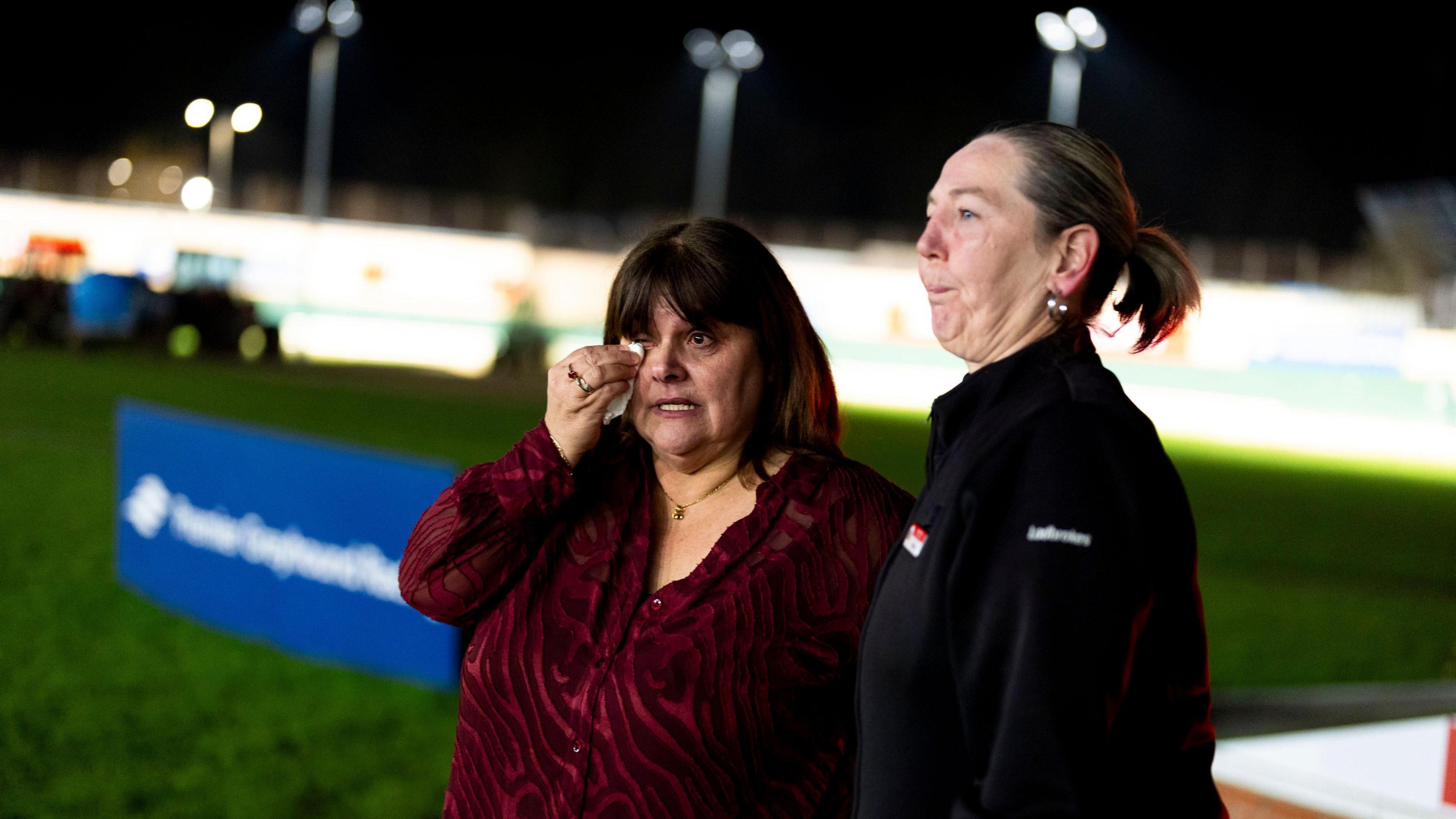 Staff cry at the Crayford Greyhound Stadium in London, after the final race is held. In the background you can see blurred sponsorship hoardings and the lights illuminating the track