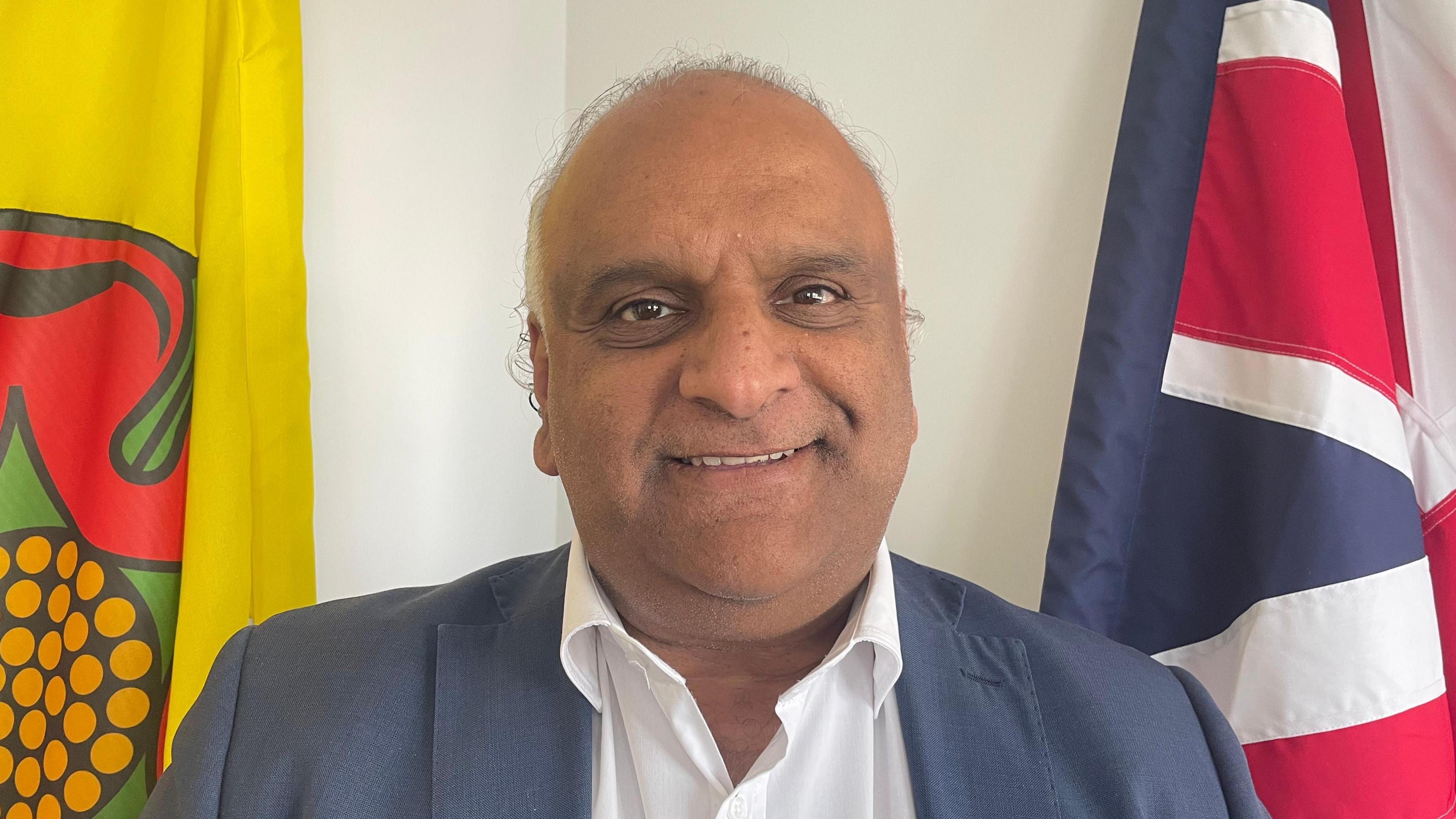 County Councillor Azhar Ali is wearing a blue suit with un unbuttoned white collared shirt. To his left is the Lancashire flag, to his right, the Union flag.