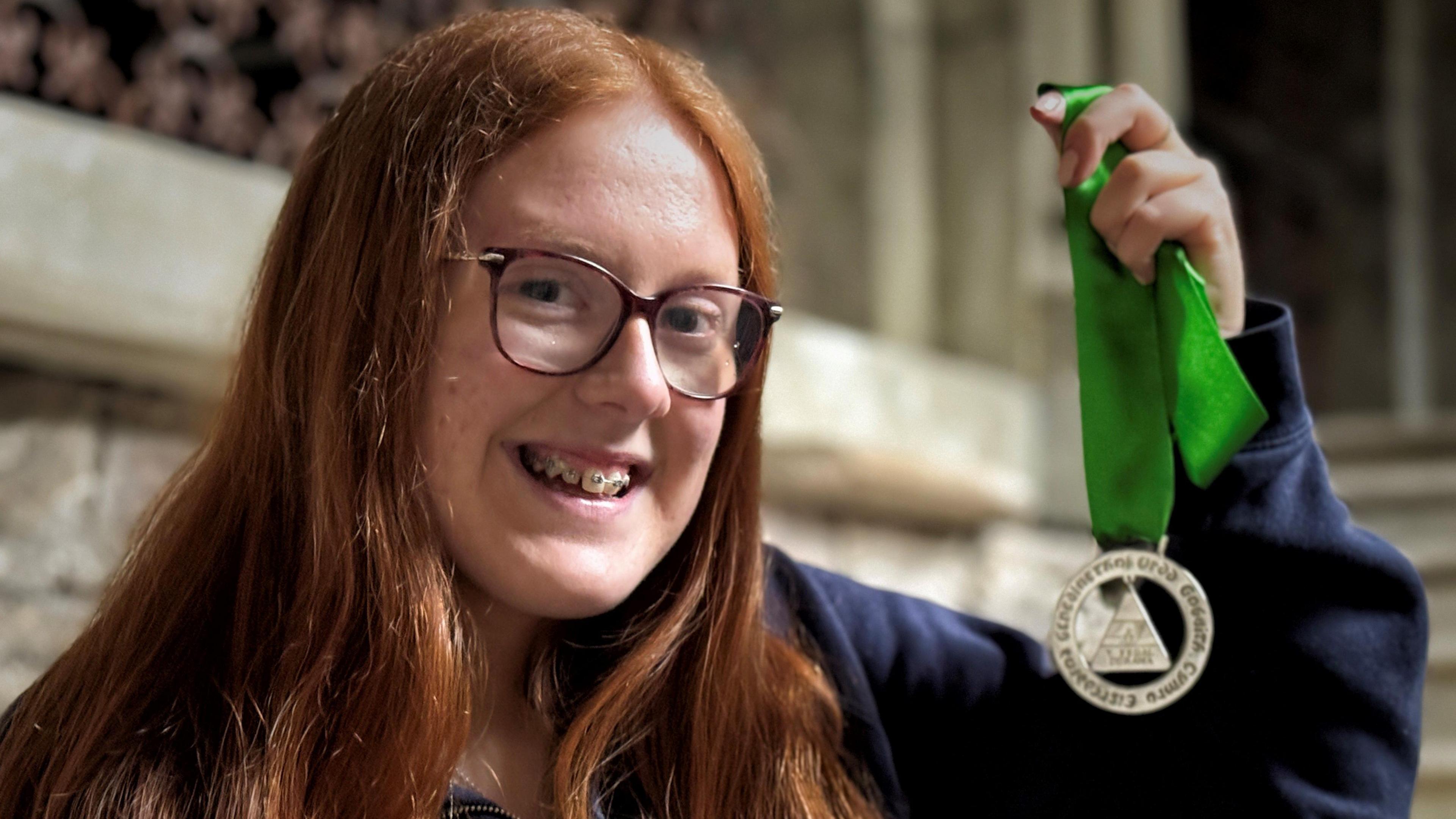 Elin Undeg Williams holding medal with green ribbon and smiling. She has long red hair, wears glasses and has braces and wears a navy top