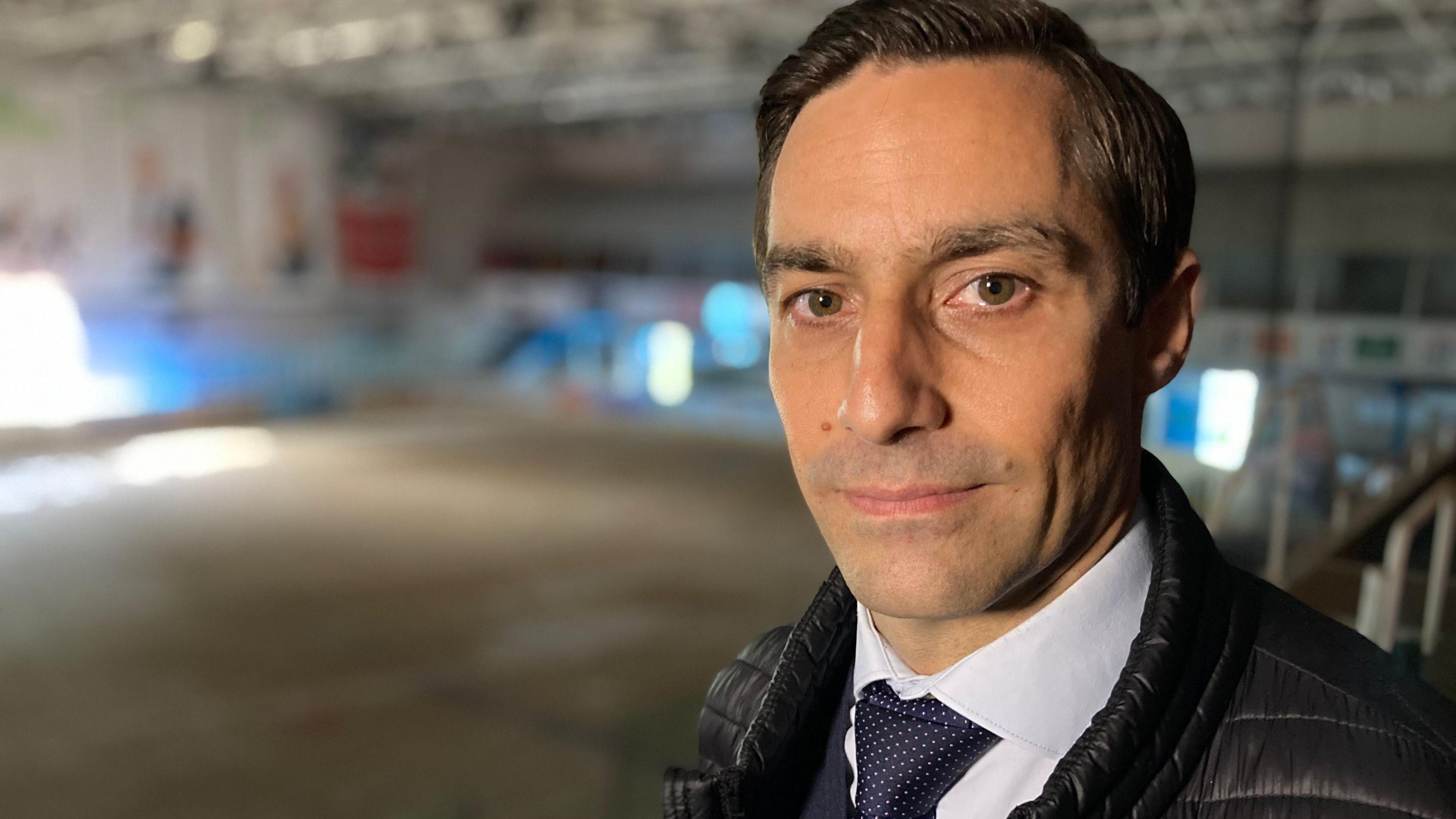 Barnaby Rich stands in the gallery of the ruined Link Centre ice rink, which is out of focus in the background. He has dark brown hair and is wearing a shirt, tie and a down-type filled jacket