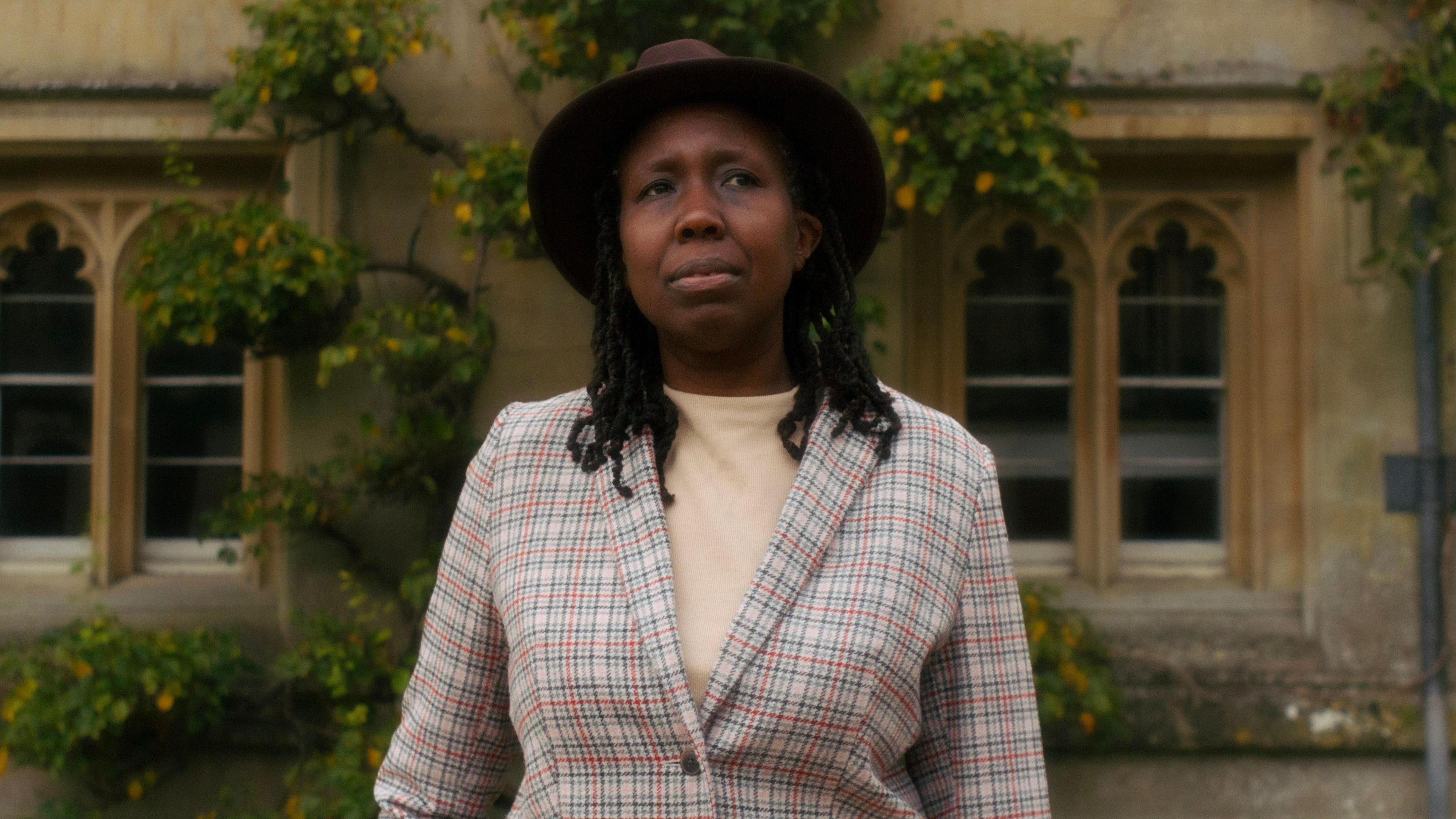 A headshot of Prof JC Niala. She is wearing a brown hat. Trees are growing by the building behind her.