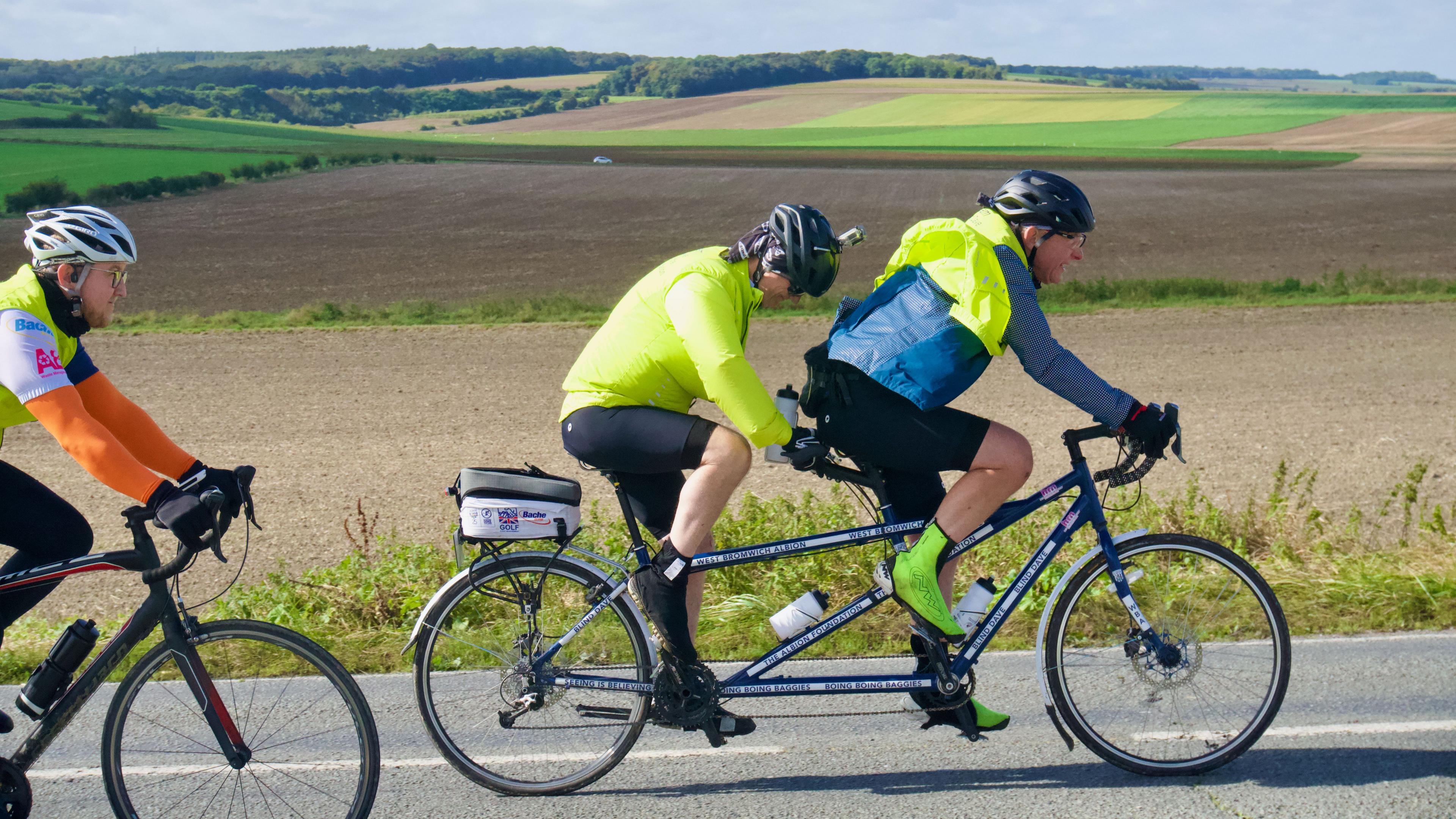 Two men on a tandem bicycle with a fellow cyclist behind