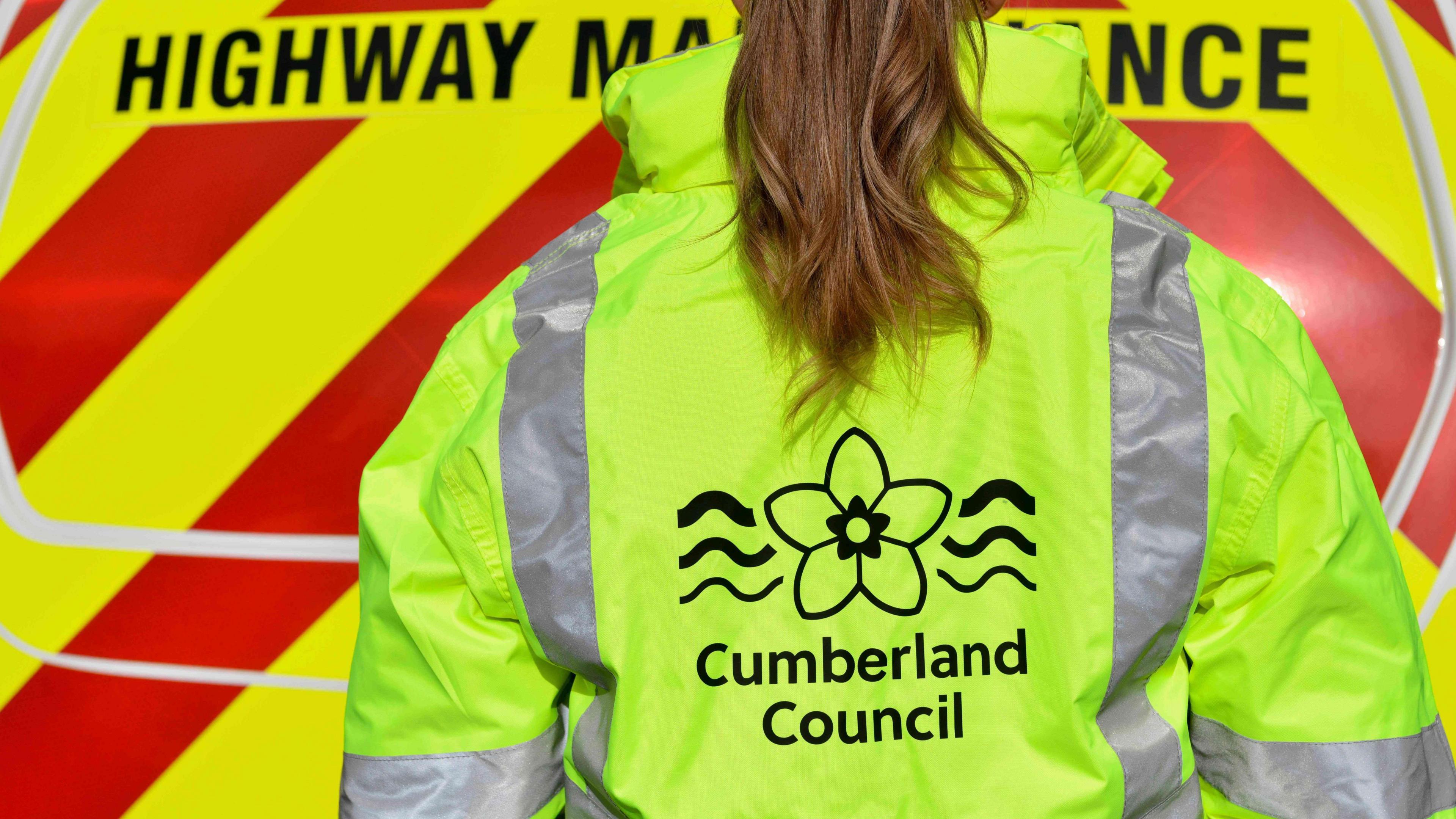 A woman wearing a high-vis jacket with the words 'Cumberland Council' on the back faces the rear of a highway maintenance van