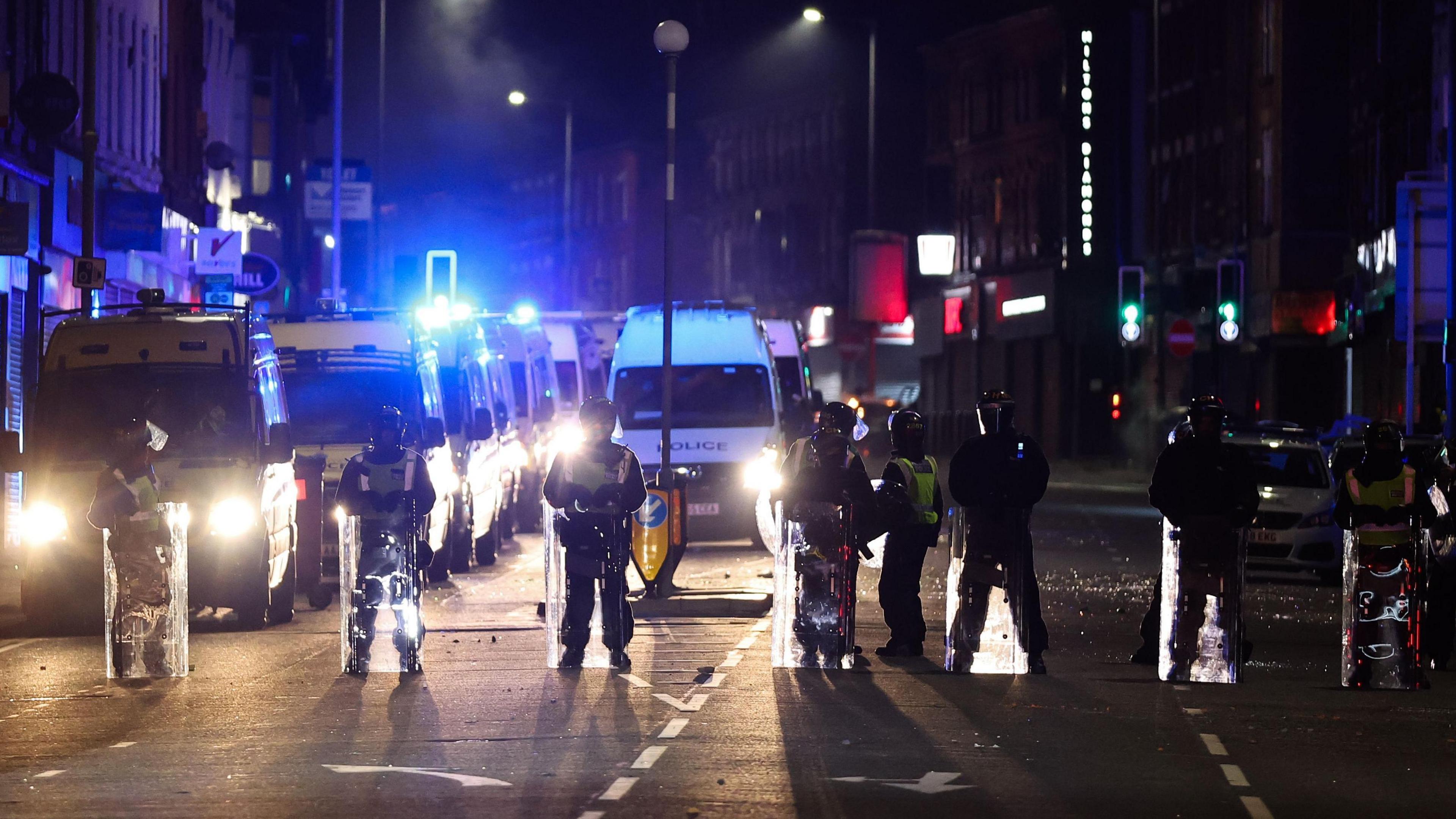 Picture shows riot police with plastic shields standing in a line in the dark with a number of emergency services vans parked behind them with their headlights on. They are stopping traffic and pedestrians passing through County Road in Walton on 3 August 2024.