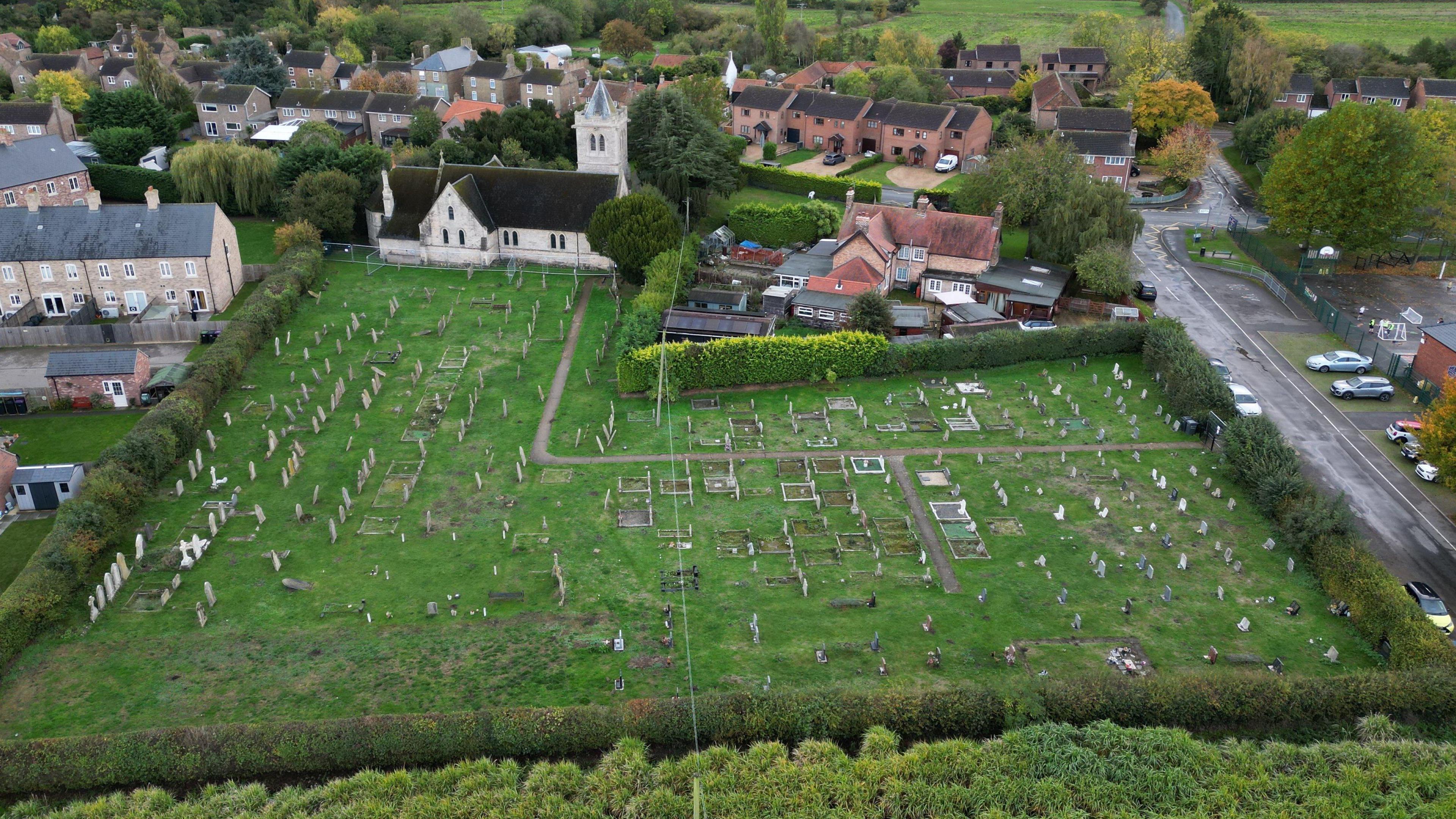 An aerial shot shows the entire graveyard. There are older gravestones towards the top of the picture where the church is, and empty plots to the bottom of the cemetery. There is a hedge around the graveyard, and no obvious access to the newer graves at the bottom of the picture. Both the grass and cemetery are green in colour, and their are homes to the left and top of the cemetery's boundaries.