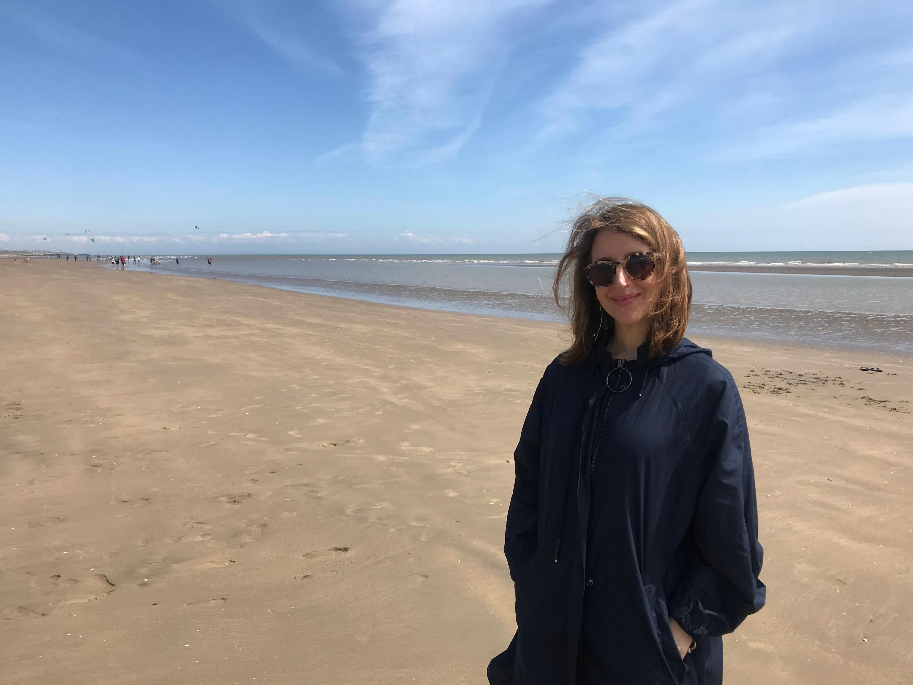 A woman with shoulder length brown hair is smiling and standing on a beach. She is wearing a black jacket and sunglasses. There is a wide expanse of sand and people can be seen in the far distance. 