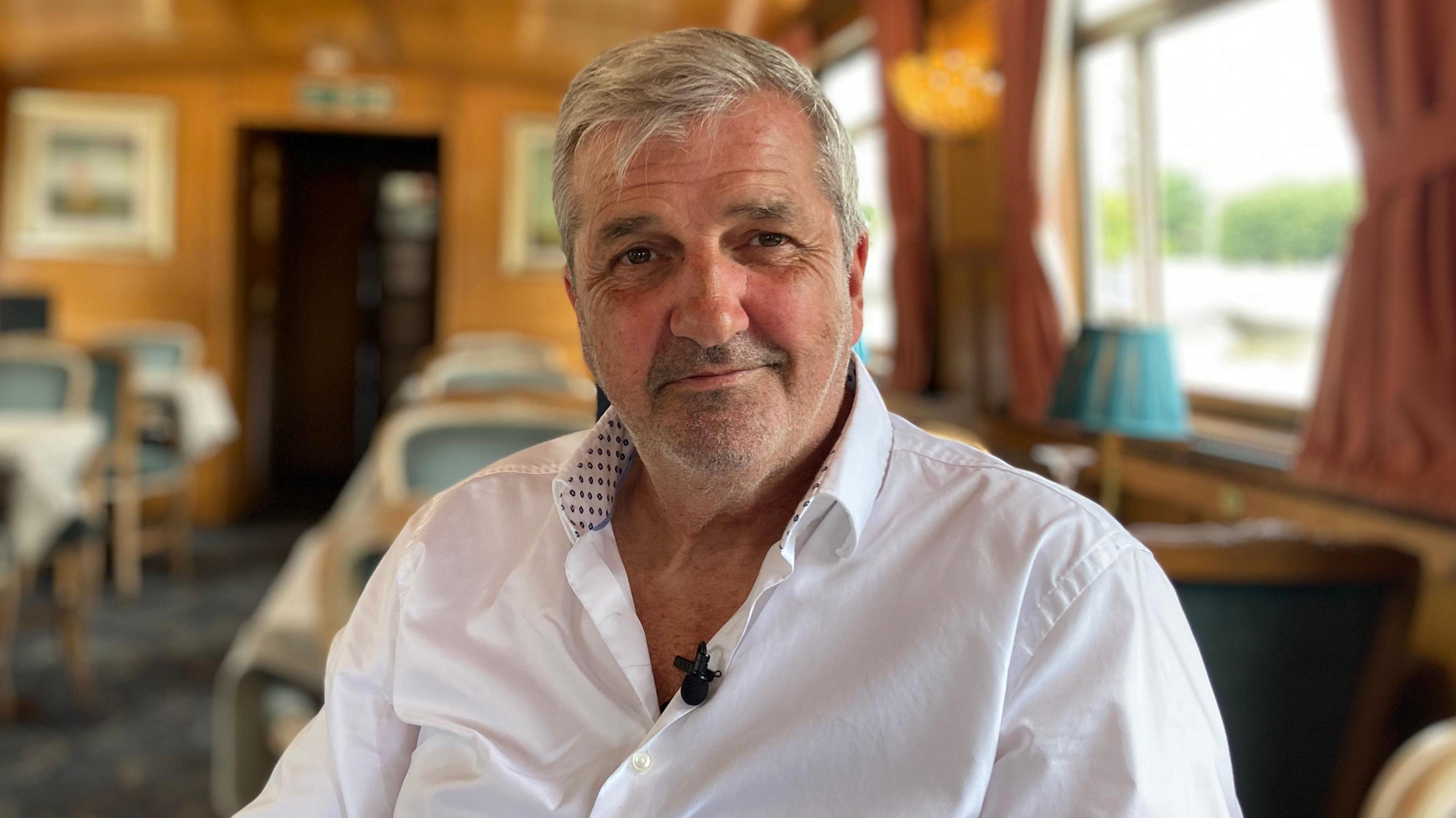 A white man with grey hair and stubble smiles toward the camera , he is sitting on a chair inside a room with wood panelling and tables and chairs behind him.