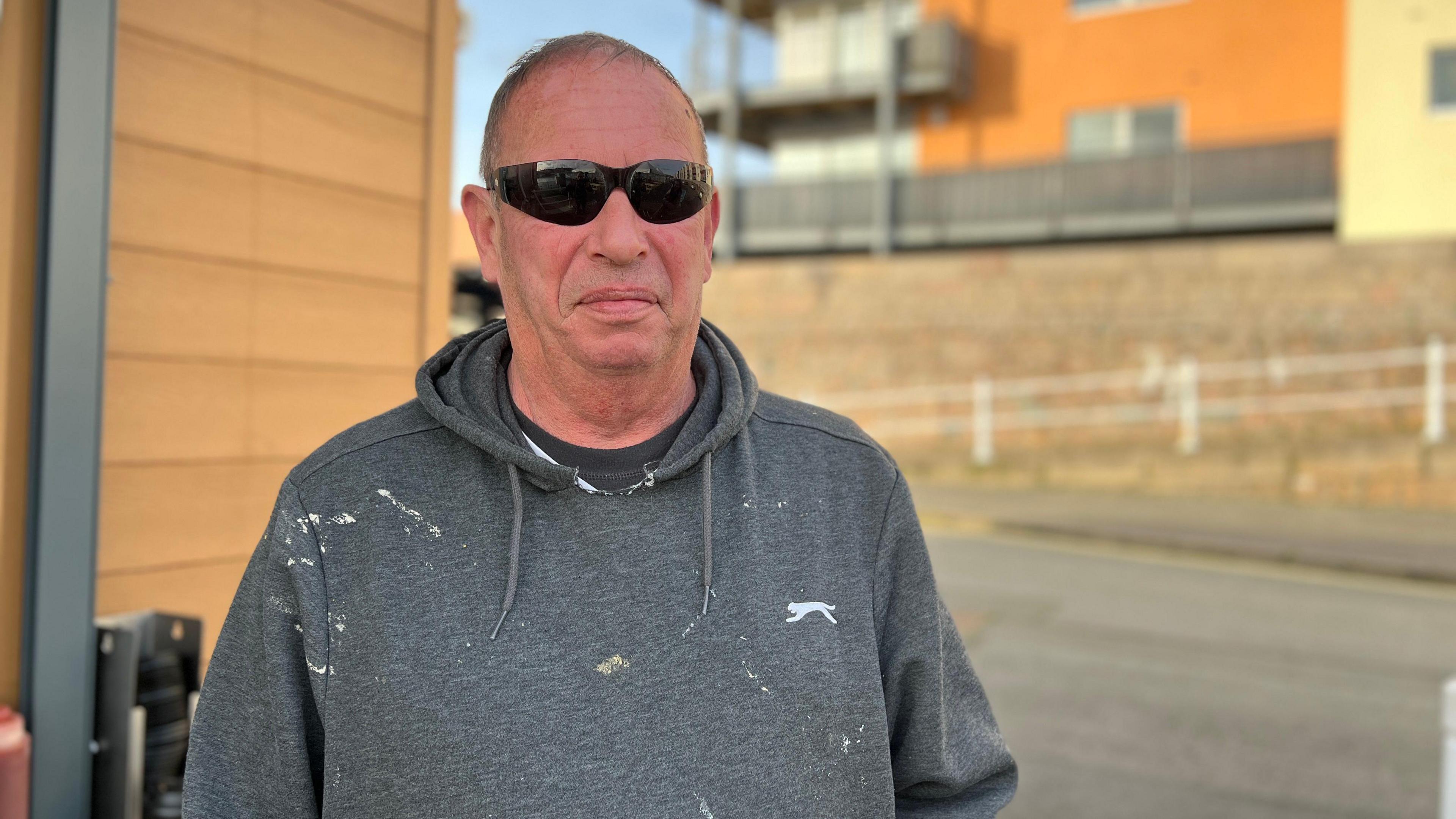 A short-haired man in a paint-splattered grey hoodie and sun glasses stands in front of some modern residential apartments.