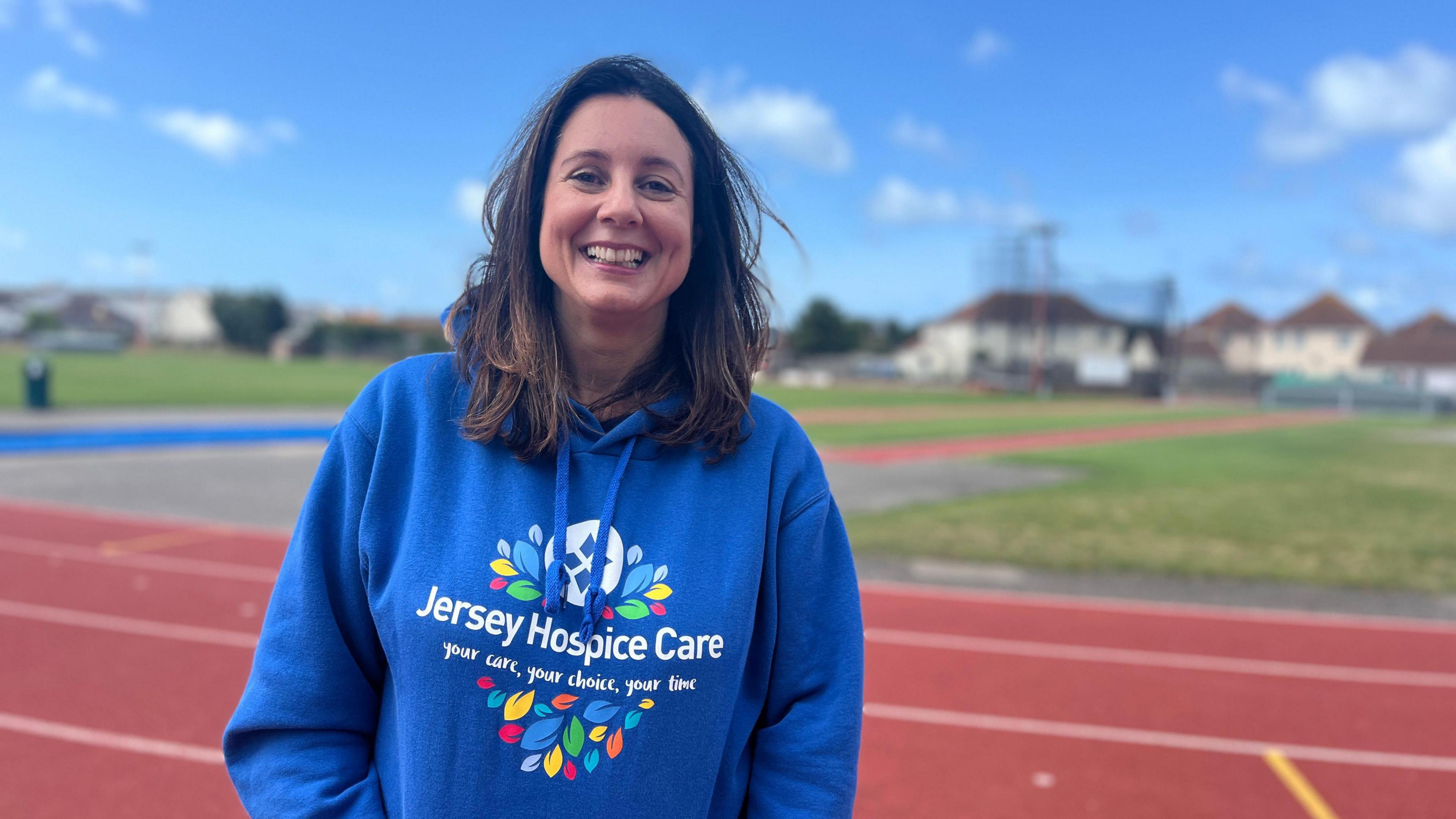 Cheri Strudwick wearing a blue Jersey Hospice Care while stood on an athletics track. She is smiling at the camera.