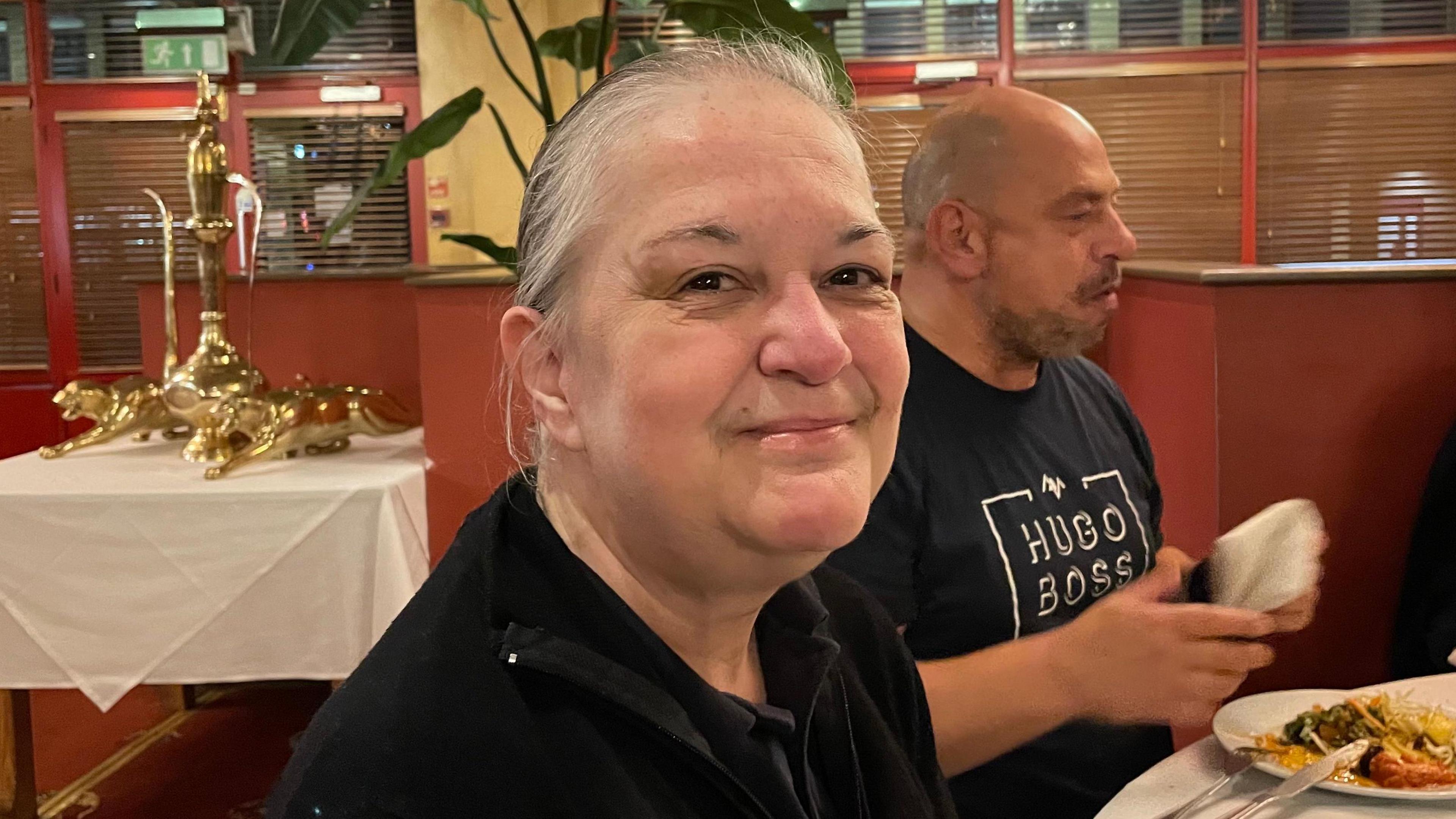 Janet Wilkinson, smiling and looking direct to camera. She is seated and wearing a black top. She is in a restaurant. There are large ornamental golden cats behind her on the left of the image and a plate of food can be seen on the right, in front of a man sitting next to her. She has dark eyes and  grey hair. 