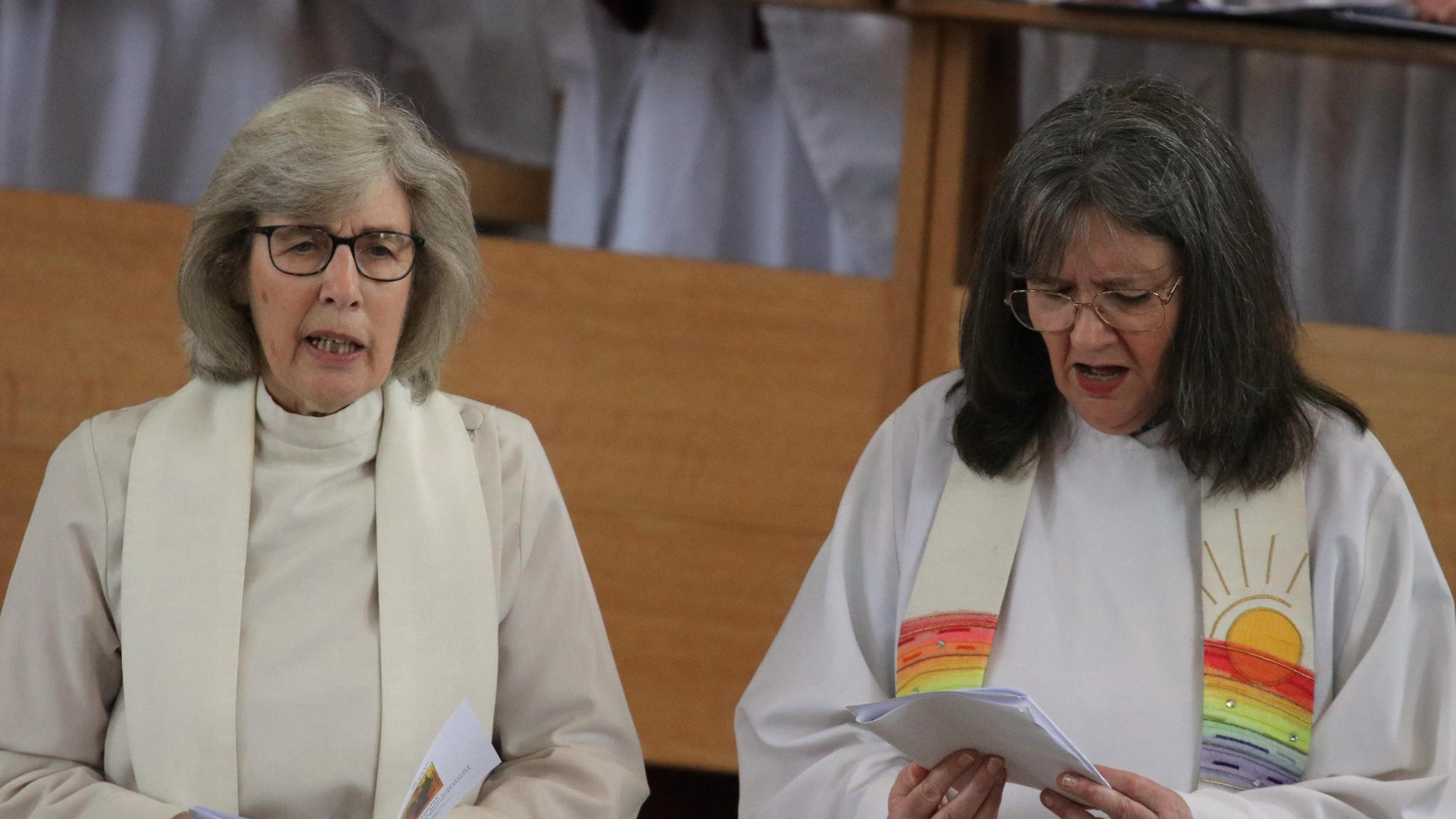 A lady with grey hair and glasses is on the left reading from a sheet of paper, whilst another lady with black hair and glasses to the right also reads from paper