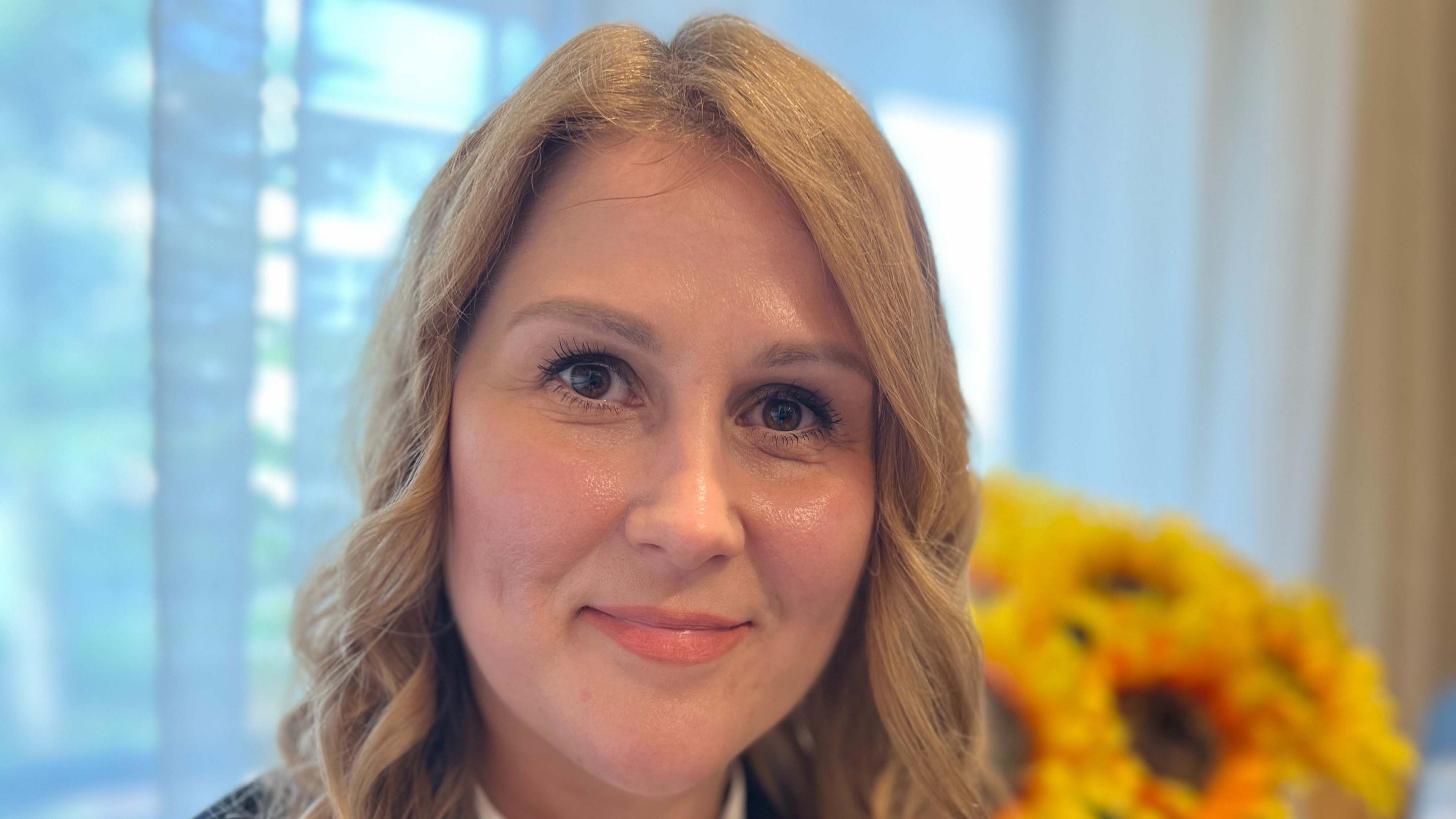 Iryna Maracine, a Ukrainian refugee, smiling straight at the camera. She has blonde shoulder length hair, and is sitting in an office with a bunch of sunflowers in the background behind her.