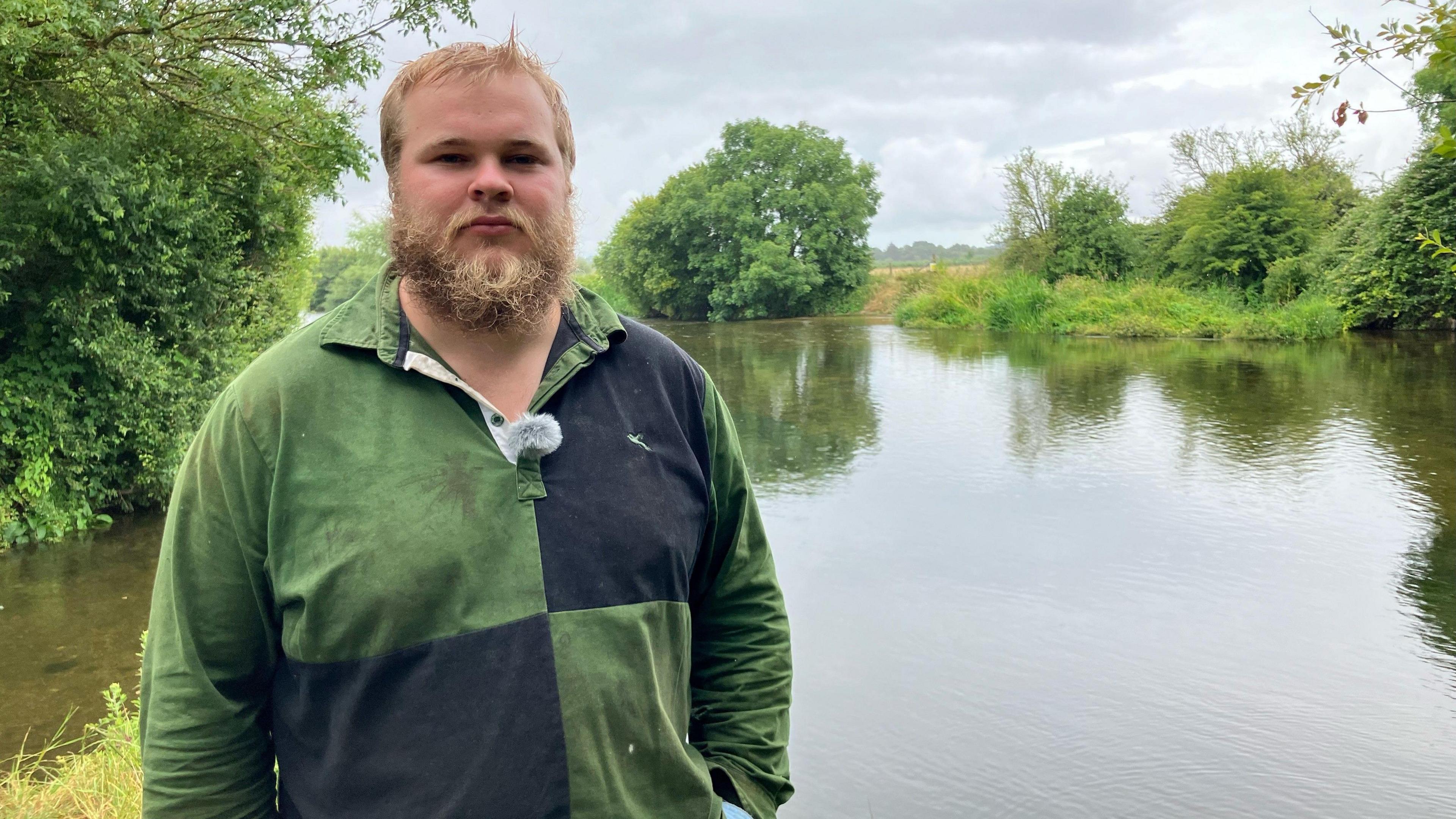 Hector Sainsbury wearing a moss green and black sweatshirt. He has a full blond beard and short blond hair and is standing on a picturesque river bank surrounded by trees