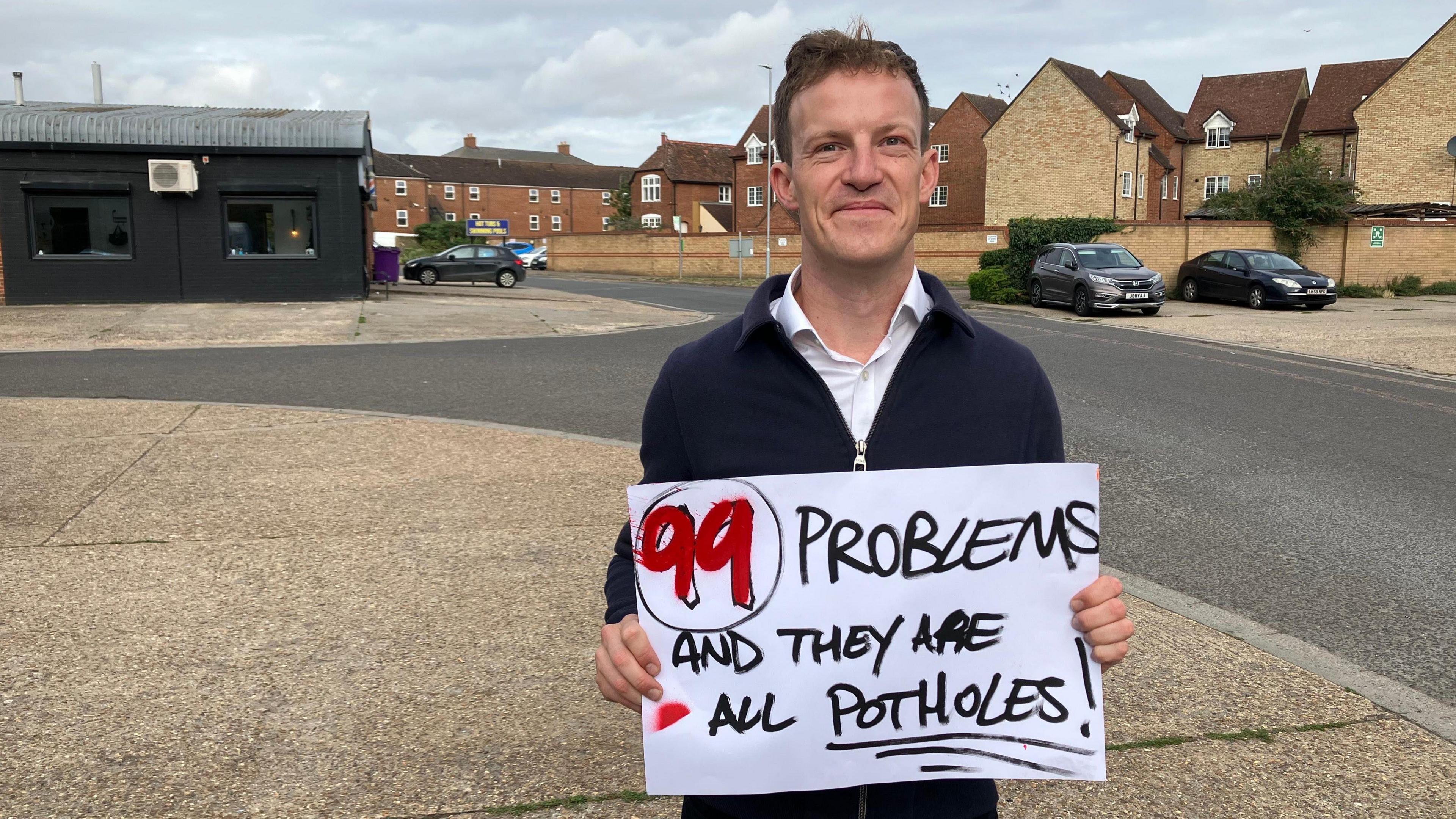 A picture of Alistair Strathern, the Labour MP for Hitchin. He is standing on an empty street, but has a protest banner in his hands that says "99 problems and they are all potholes". He is smiling and is wearing a dark blue jacket.
