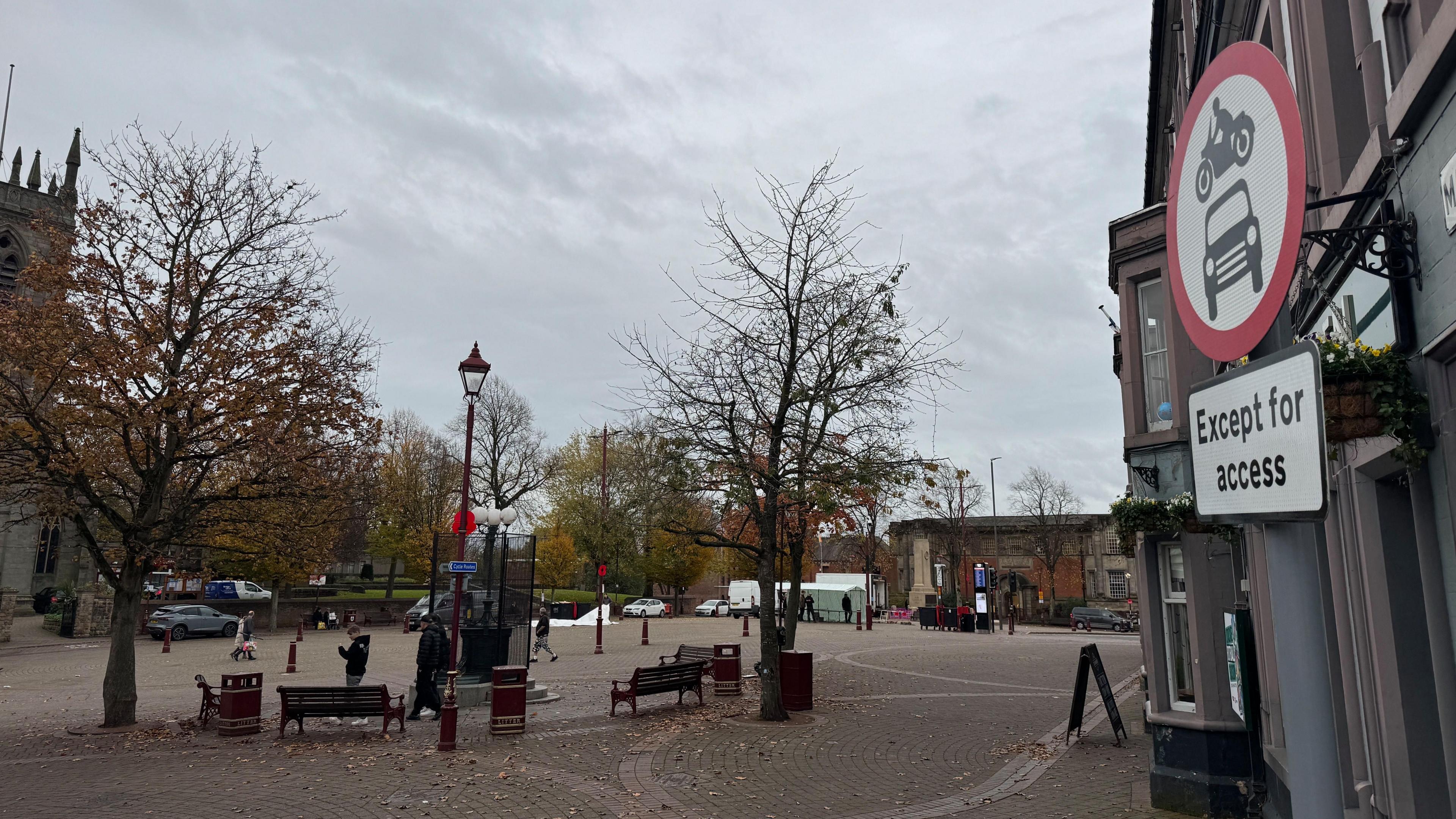 A traffic sign to the right says motorised traffic is blocked "except for access". In front is a market square with benches, trees and stalls in the distance being taken down.