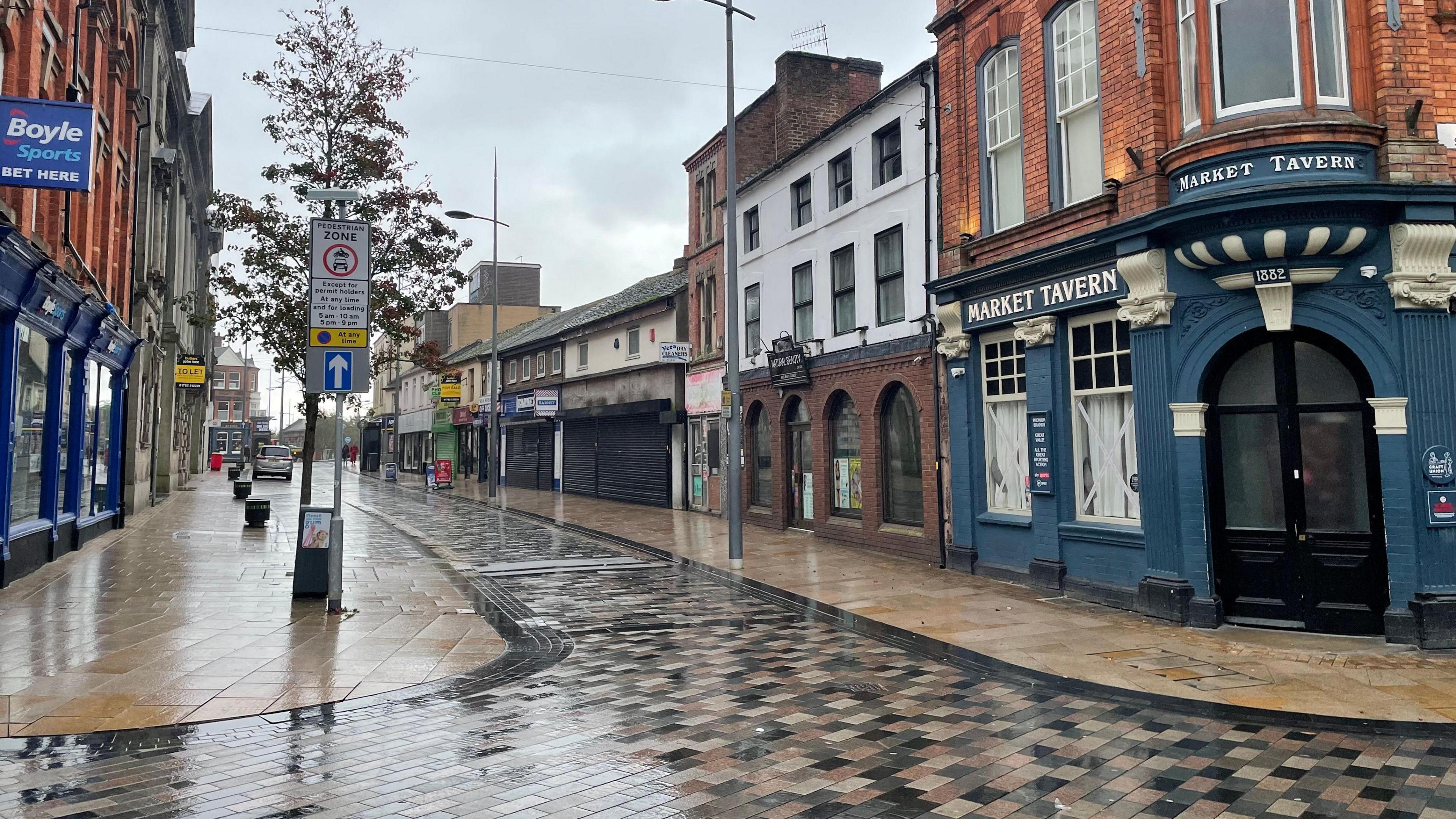 An empty street with wet paving following rain. On the right of the street on the corner is a brick building with blue panelling below reading "Market Tavern". The building has two windows and an arched doorway.