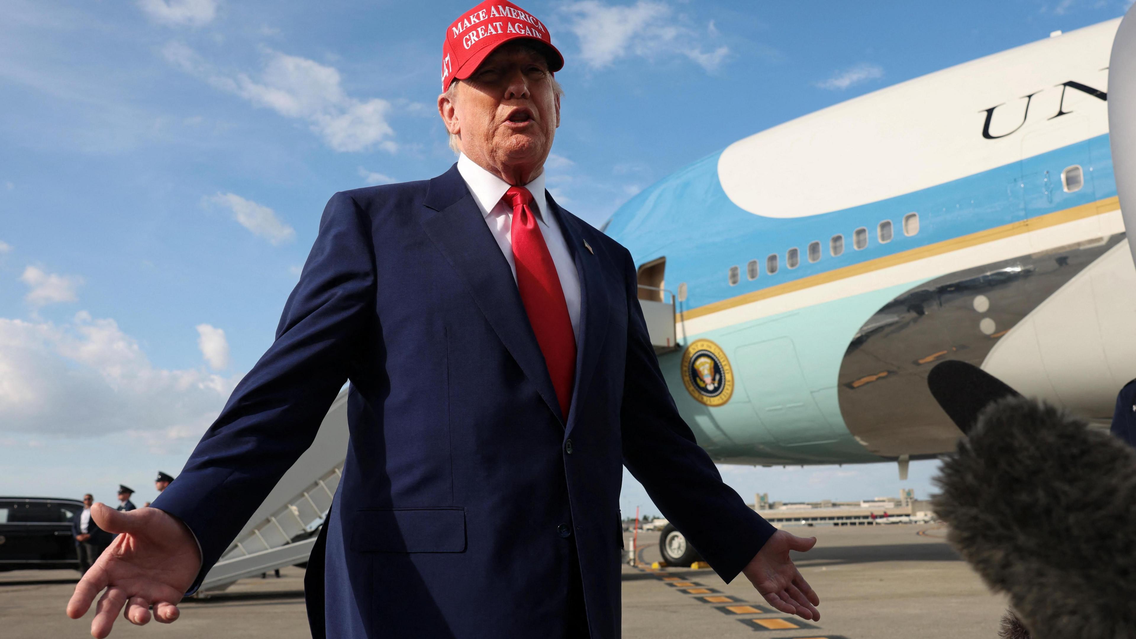 Trump in a suit and red MAGA cap talking to reporters on the tarmac at an airport on Florida on 16/2/2025