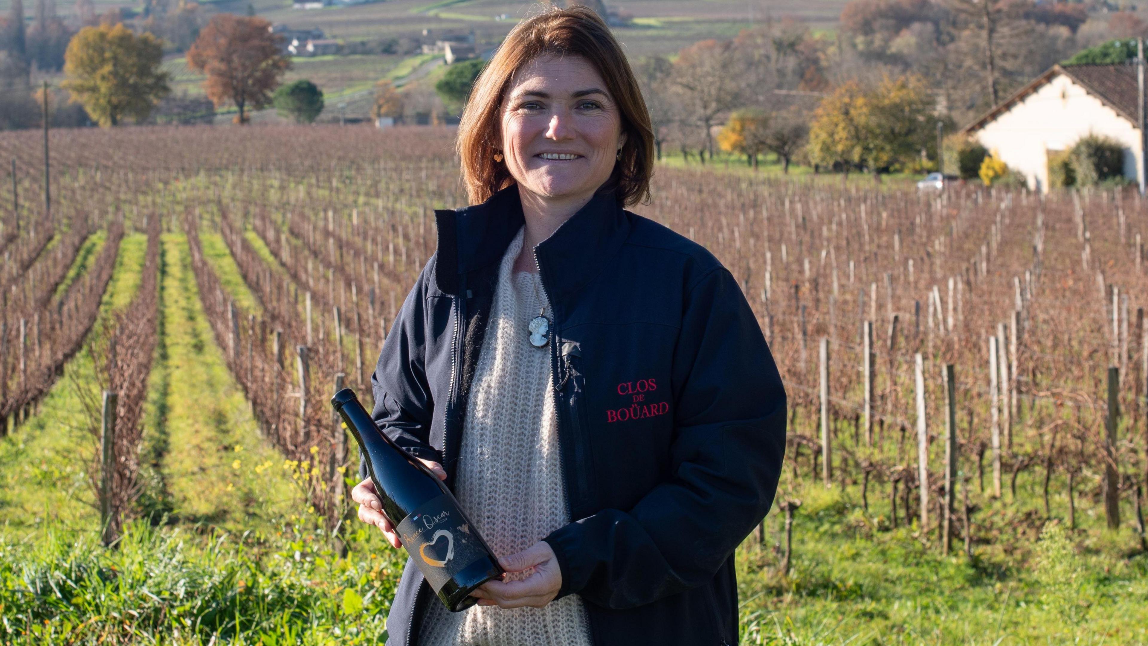 Coralie de Bouard stands in a vineyard holding a bottle of her wine