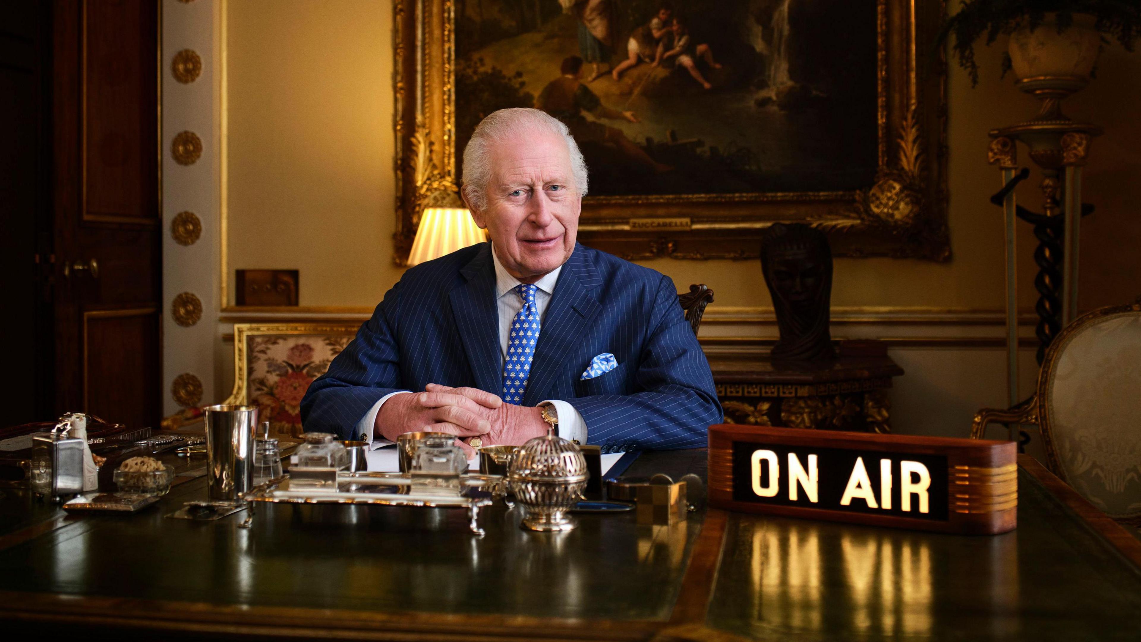 King Charles sitting at a desk with a big lit up on air sign.