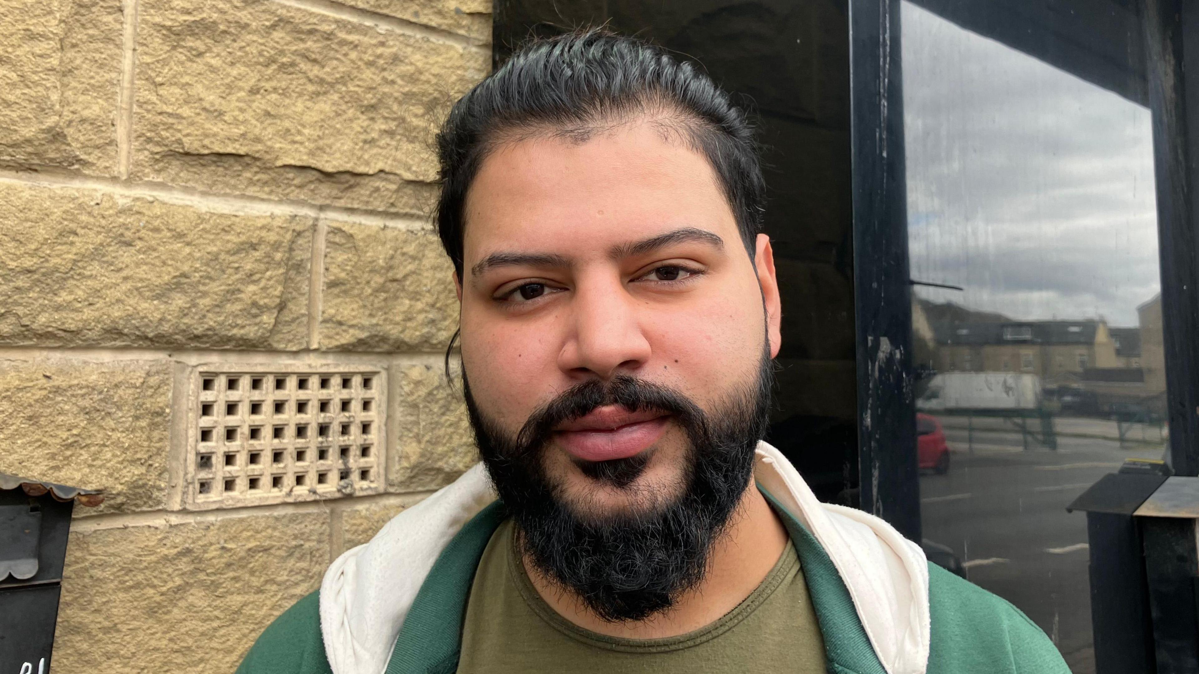 A young man with black hair and a black beard stands by a shop wall on Leeds Road in Bradford.