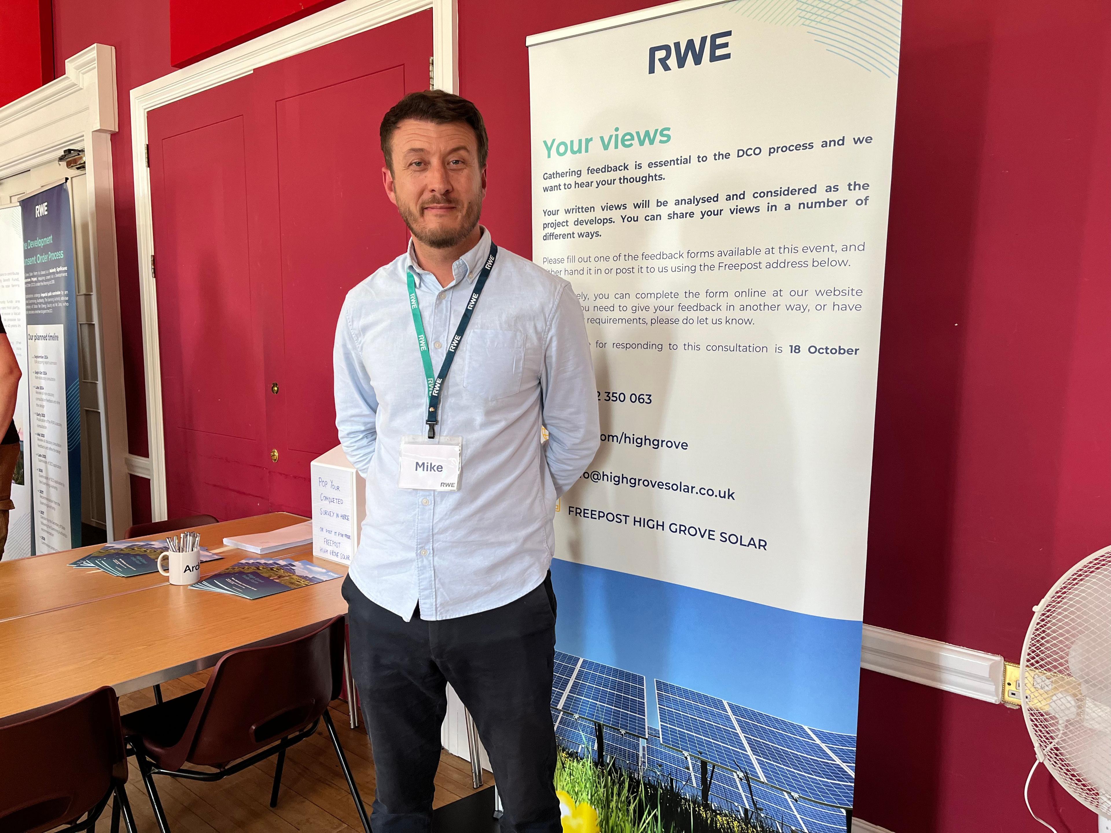 Mike Gresler from the energy firm RWE stands in front of a banner asking people to tell them what they think about the proposed solar farm.
He is wearing a blue shirt and black trousers.