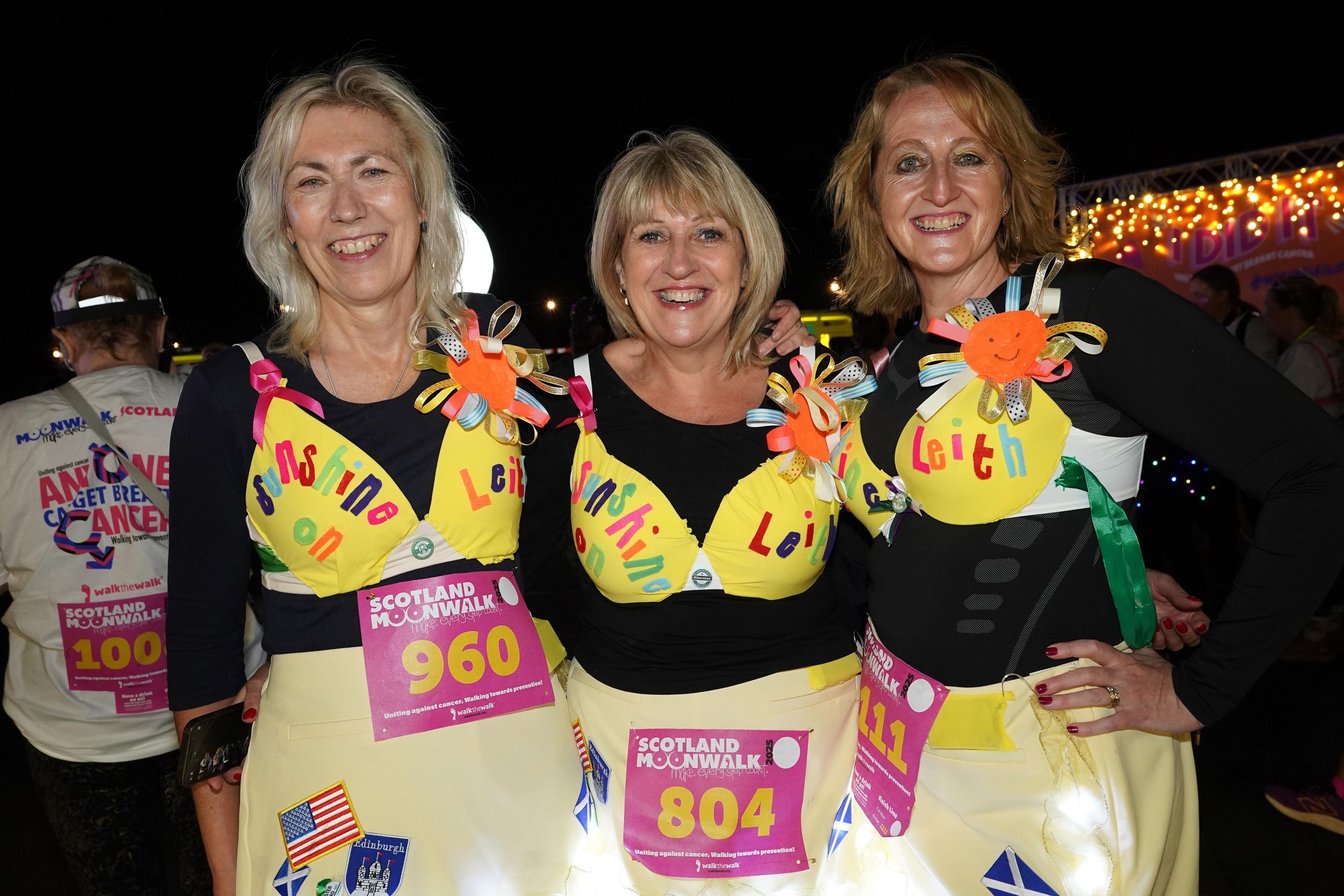 Three women in yellow bras with the words Sunshine on Leith written on top of them smile into the camera