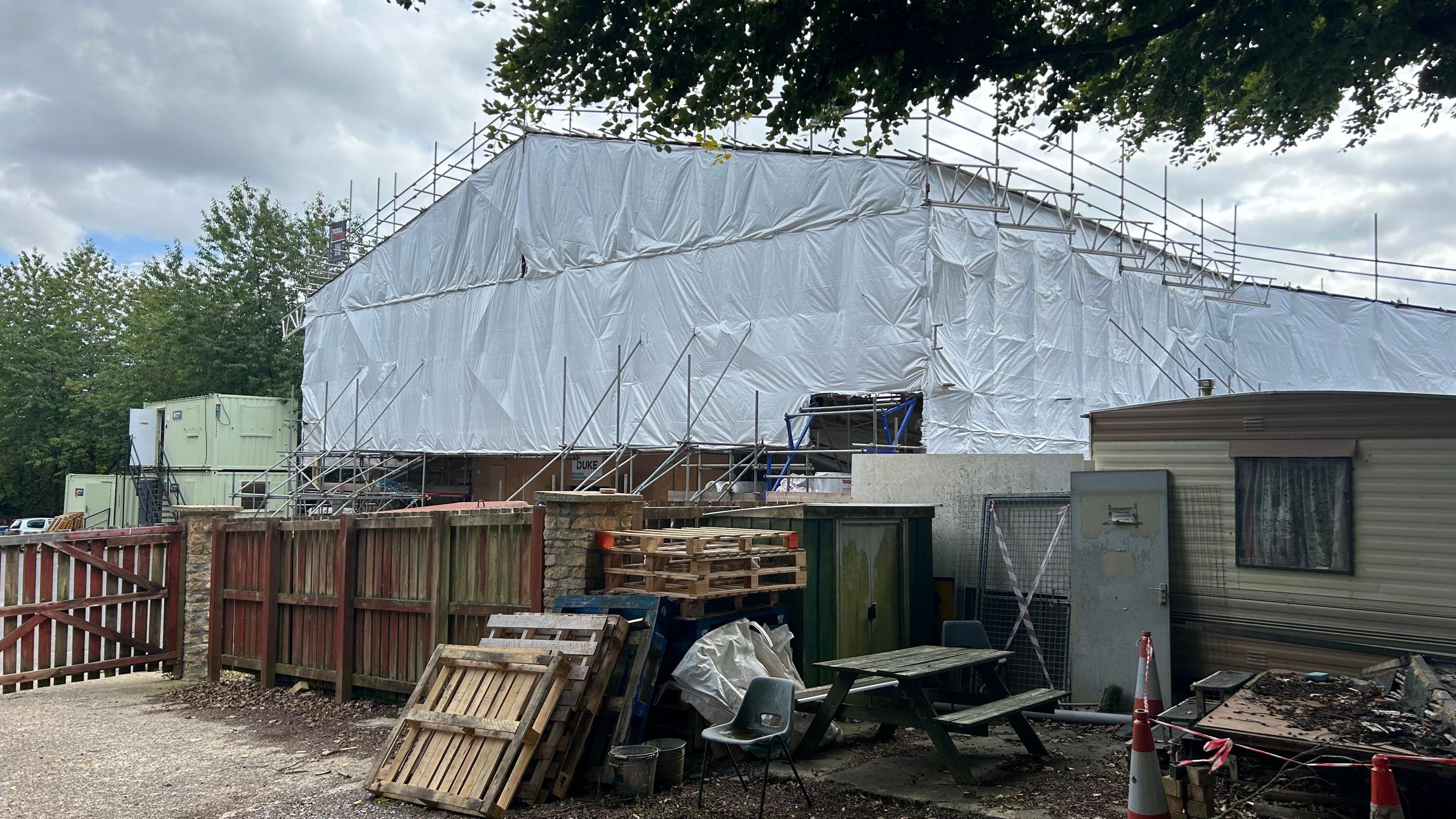 Construction work underway for the new accommodation building at Bath Cats and Dogs Home. There is a large building covered in scaffolding and plastic sheeting, and wooden pallets stacked up to the side. 