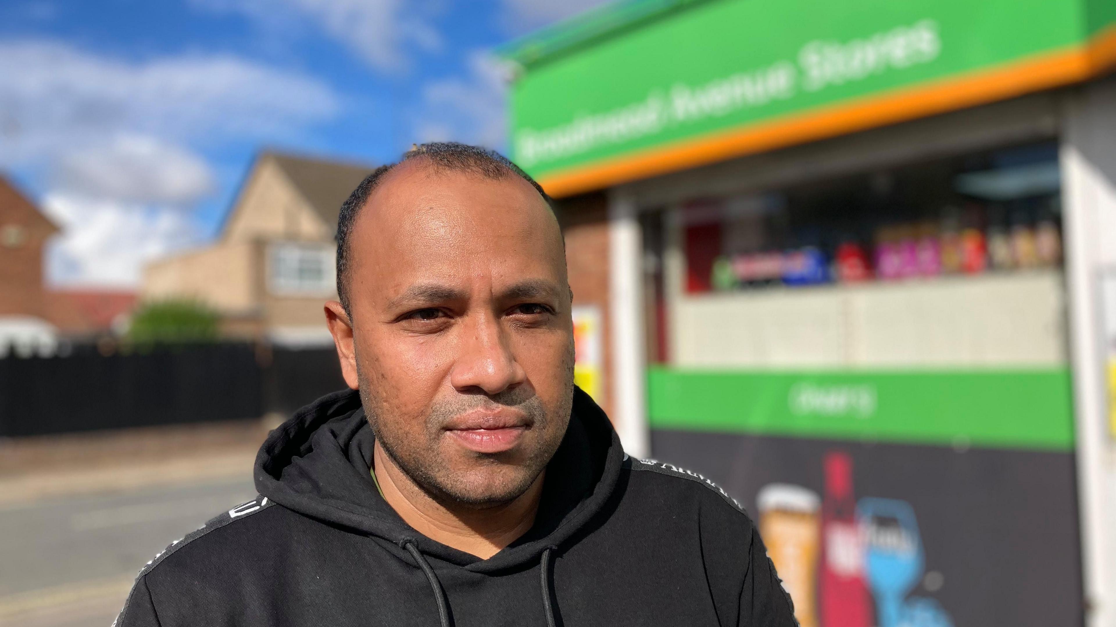 Arun Siva with very short dark hair wearing a black hoodie outside a corner shop with green sign above window.  A road and houses are visible behind him.