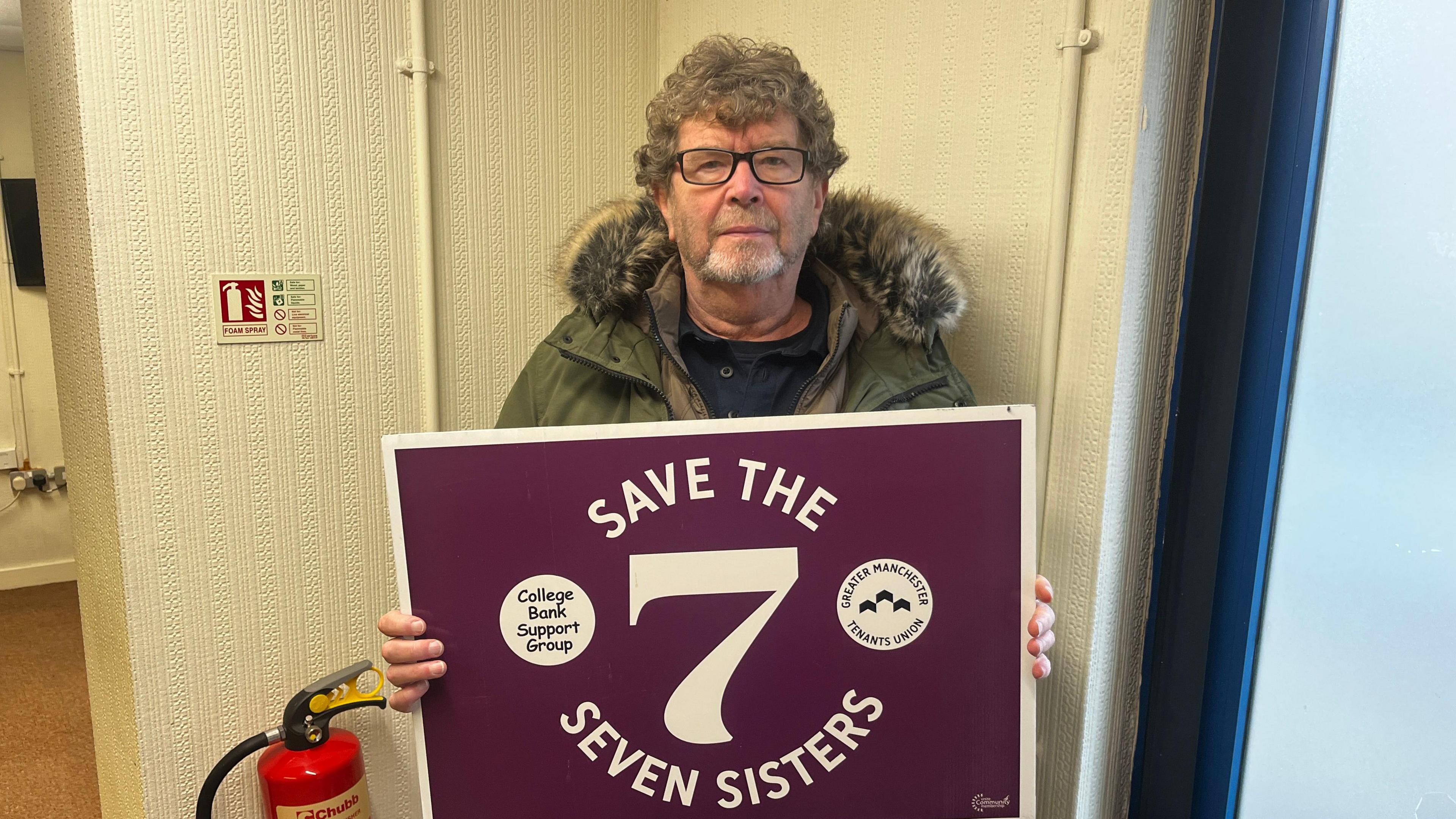 A man with grey curly hair and glasses holds a sign that reads 'save the 7 seven sisters' in a flat corridor 