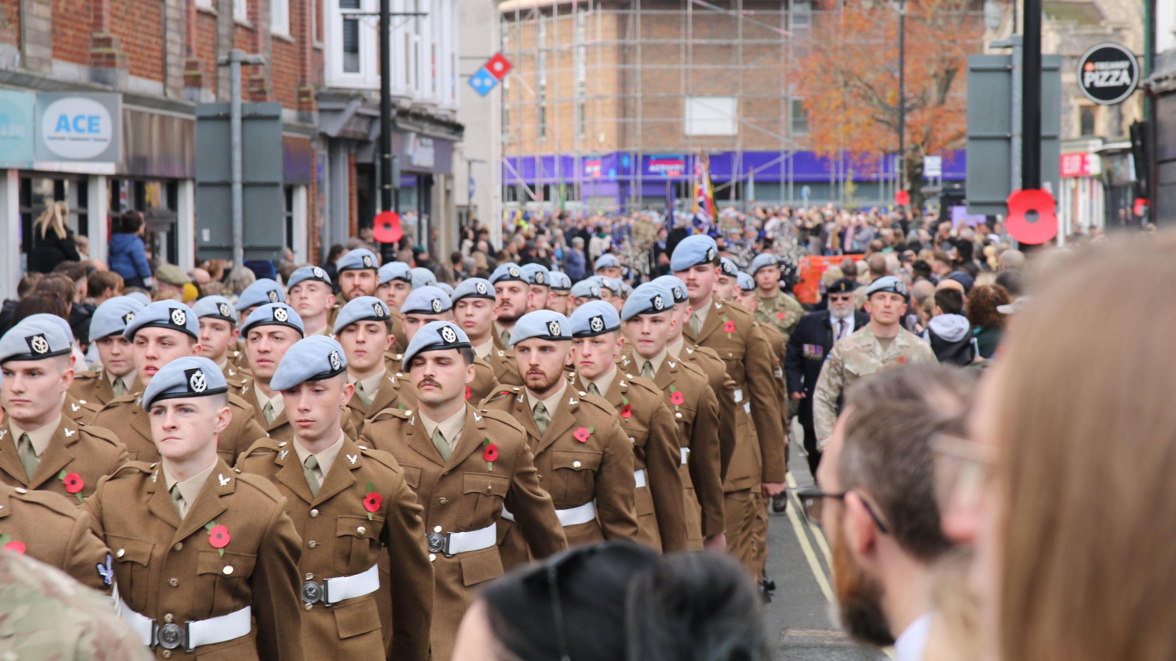 Soldiers wearing poppies marching through a street in Andover on Remembrance Sunday. People are watching them from the side. It is overcast.