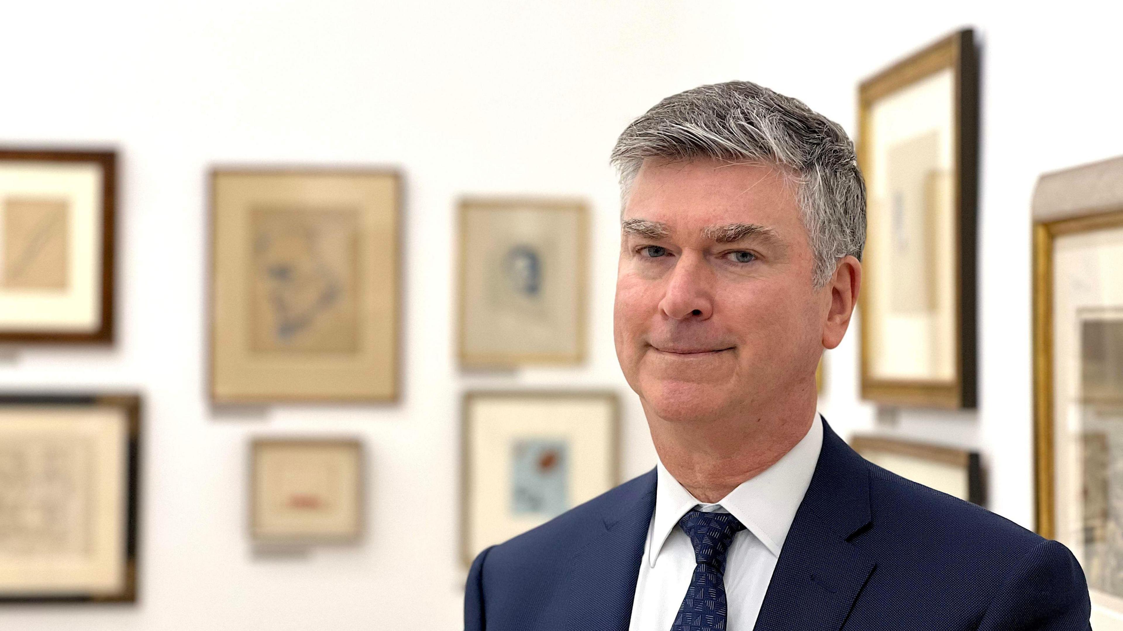 A picture of a man in a blue suit an tie. He is looking at the camera directly. He's standing in a white art gallery, and there are lots of 20th century art on the walls. They are blurry. 