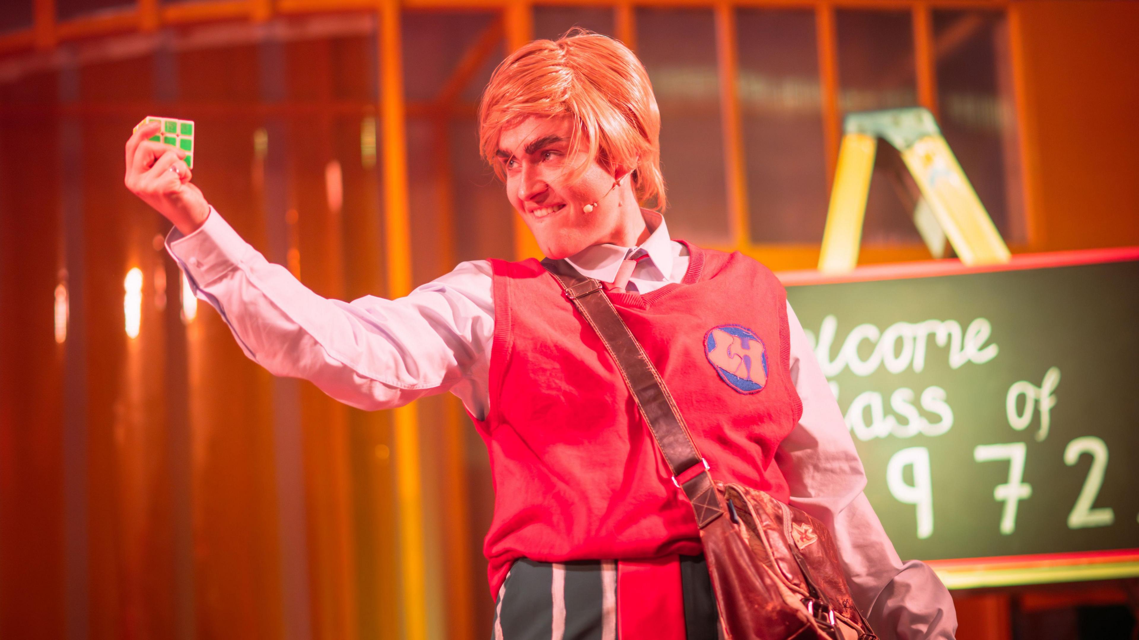 Male actor on stage facing the side of the stage and holding up a Rubiks cube. He is wearing a red tank top with the letters LH on a badge over a pink shirt. He also has stripey red, green and white trousers on and has a brown satchel over his shoulder. He is partly obscuring a green sign behind him that says Welcome Class of 1972.