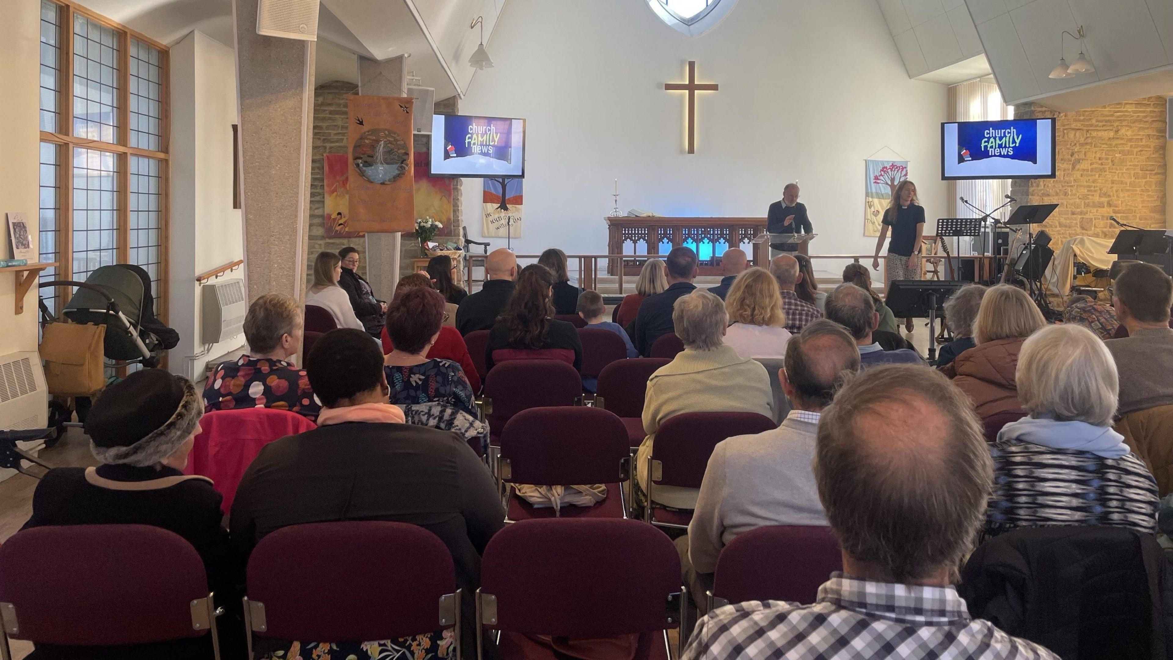 People sat on red chairs at a church while people are stood at the front talking