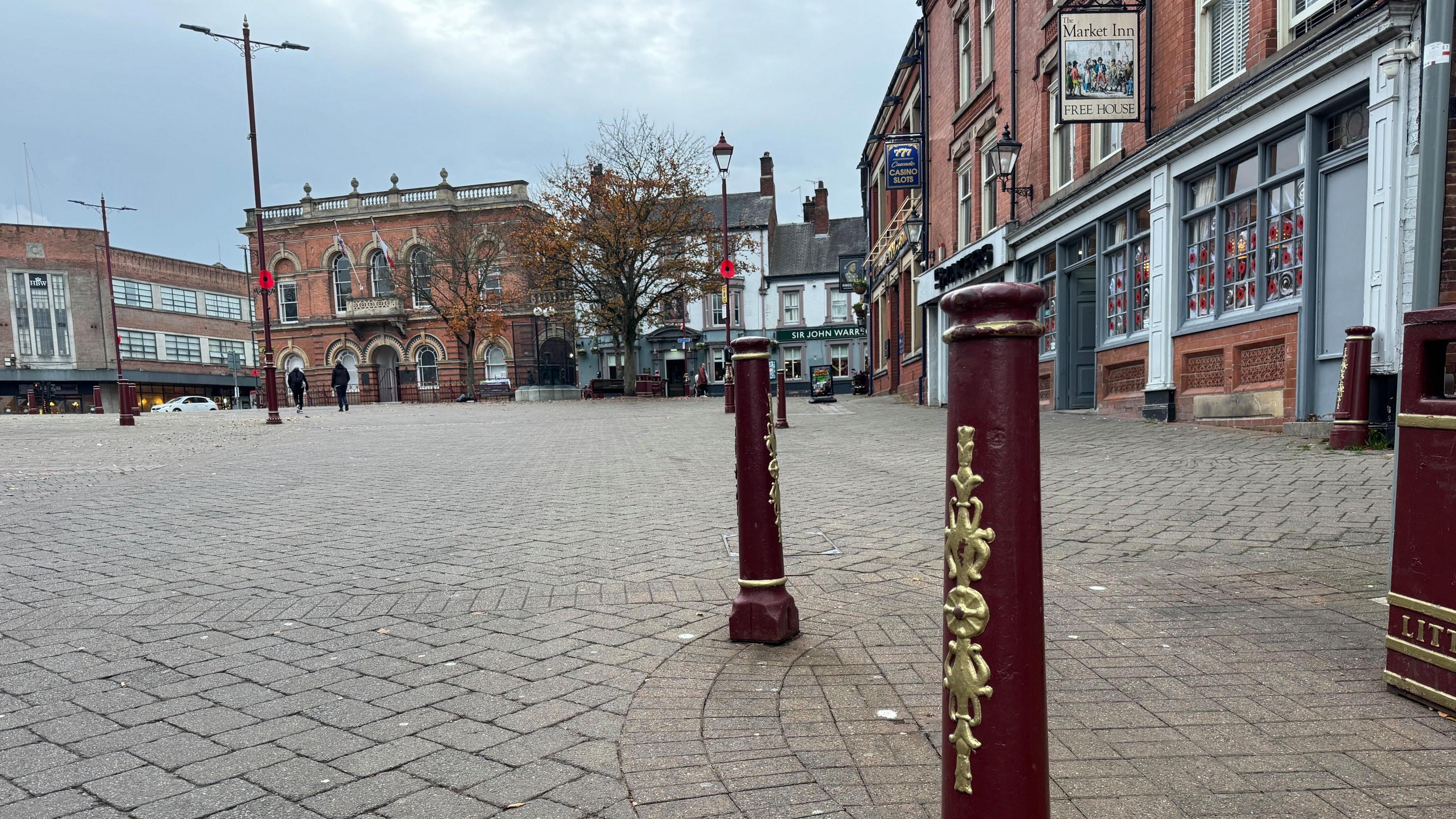 Two dark red and bollards with gold decoration protect an area to the right of the scene including the frontage of The Market Inn pub. The rest of the area opens out into Ilkeston Market Place