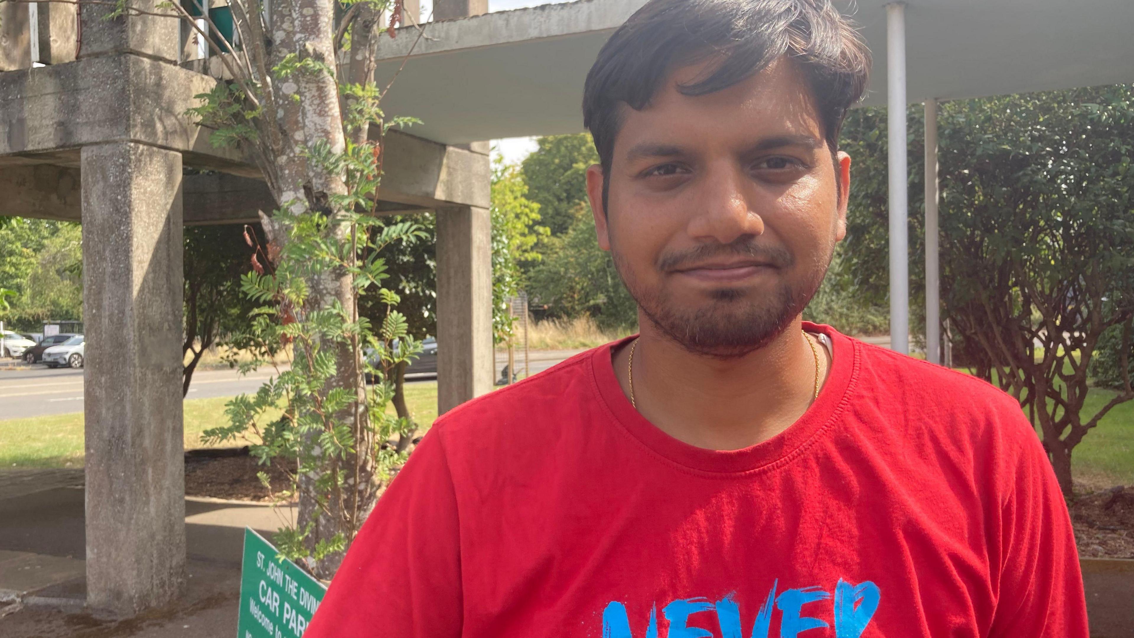 Niraj Gupta is standing outside in the sunshine with a tree behind and brick pillars him. He is wearing a red T-shirt with a blue logo.