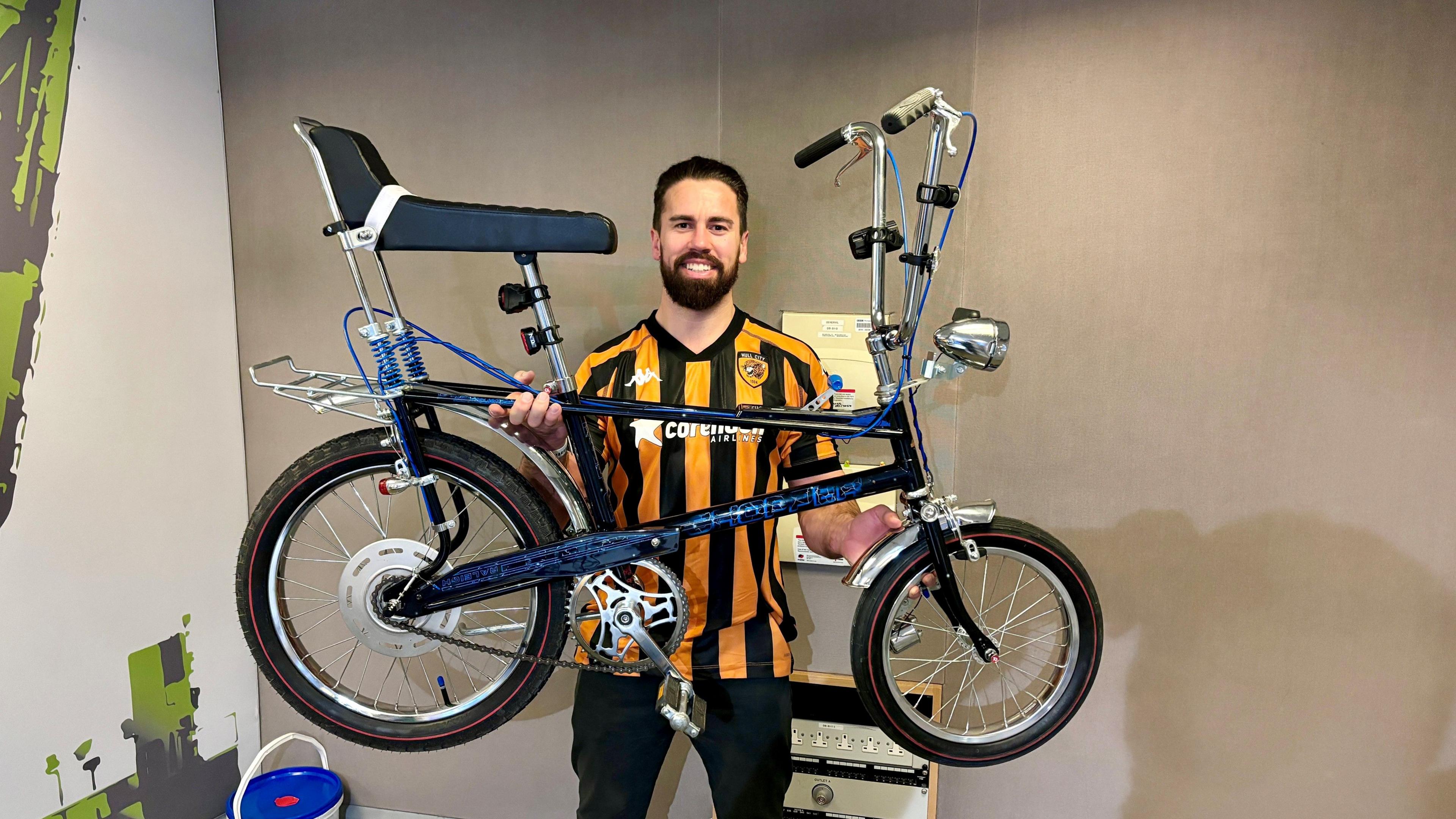 Mike Humphreys pictured wearing a Hull City FC football shirt, holding aloft his limited edition Raleigh Chopper, which has a front light and rack over the back wheel.