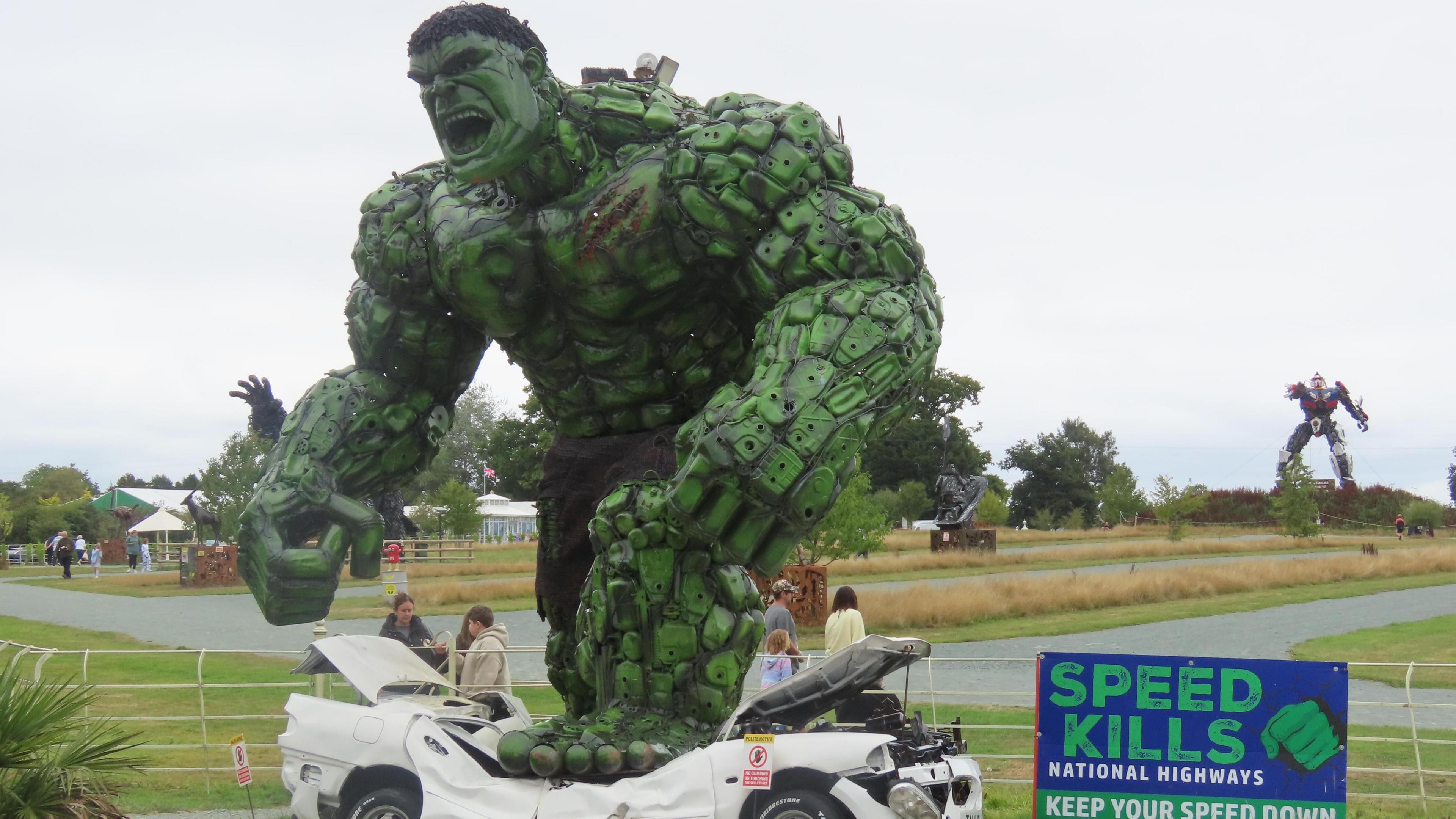 Sculpture of a giant Hulk, with its foot on a crushed car. The sculpture is next to a sign which reads Speed Kills, National Highways, Keep your speed down. 