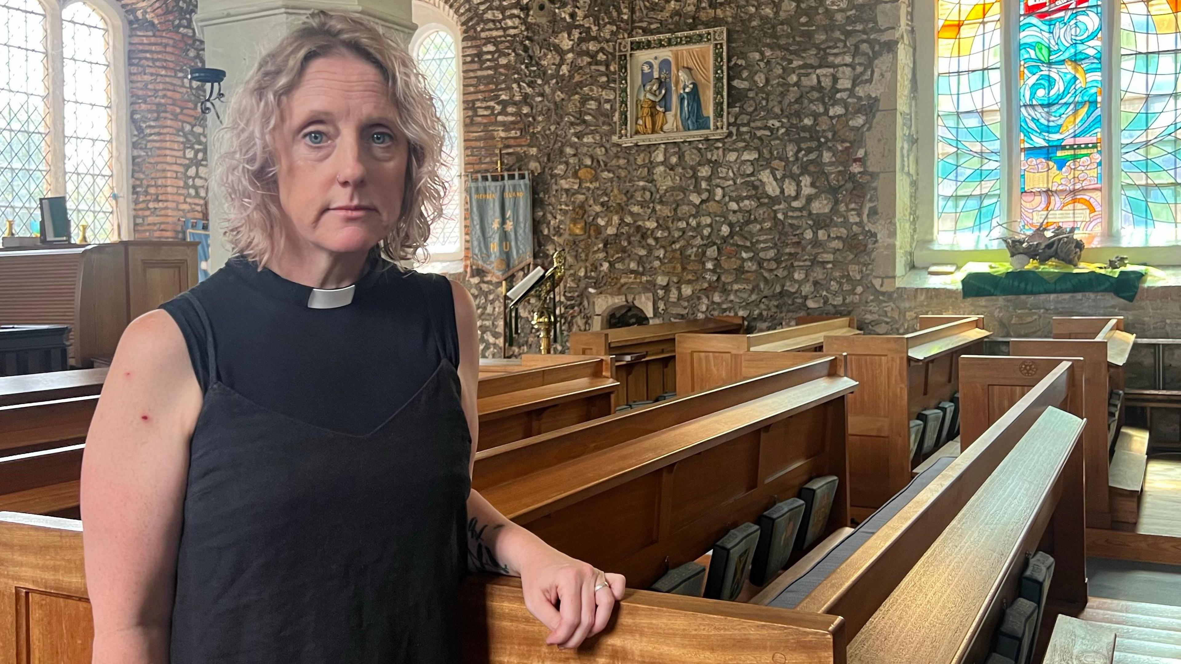 Jo Parrott wears a black top and a white dog collar, as she leans against a pew in a medieval looking church, with stone walls and stained-glass windows.
