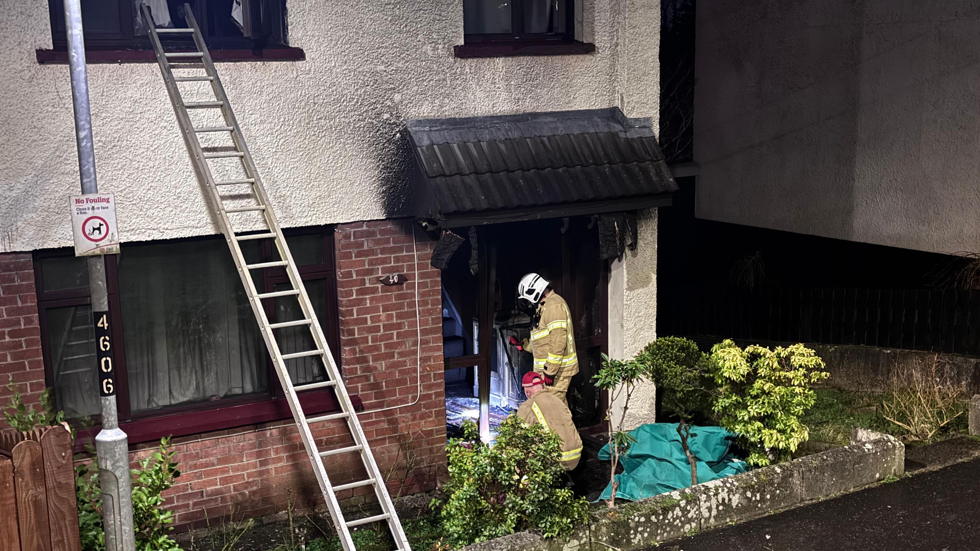 A home showing signs of smoke damage with two fire fighters standing in uniform at the door. 